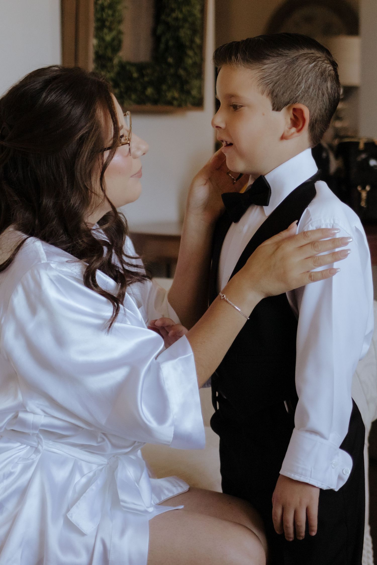 Bride in white satin robe tenderly adjusts young boy's bow tie as he stands in formal black and white attire.