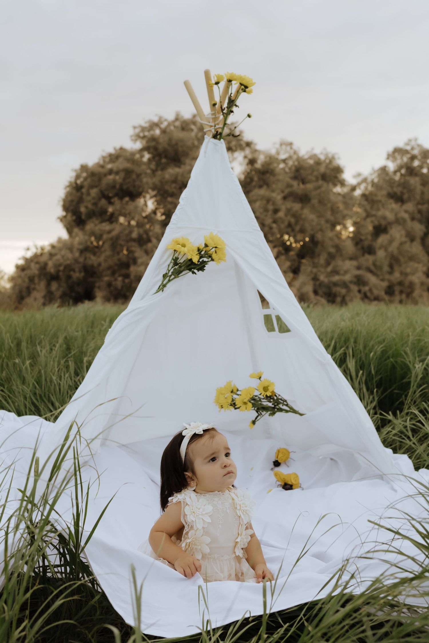 Baby girl sits in front of white teepee decorated with yellow flowers in field.