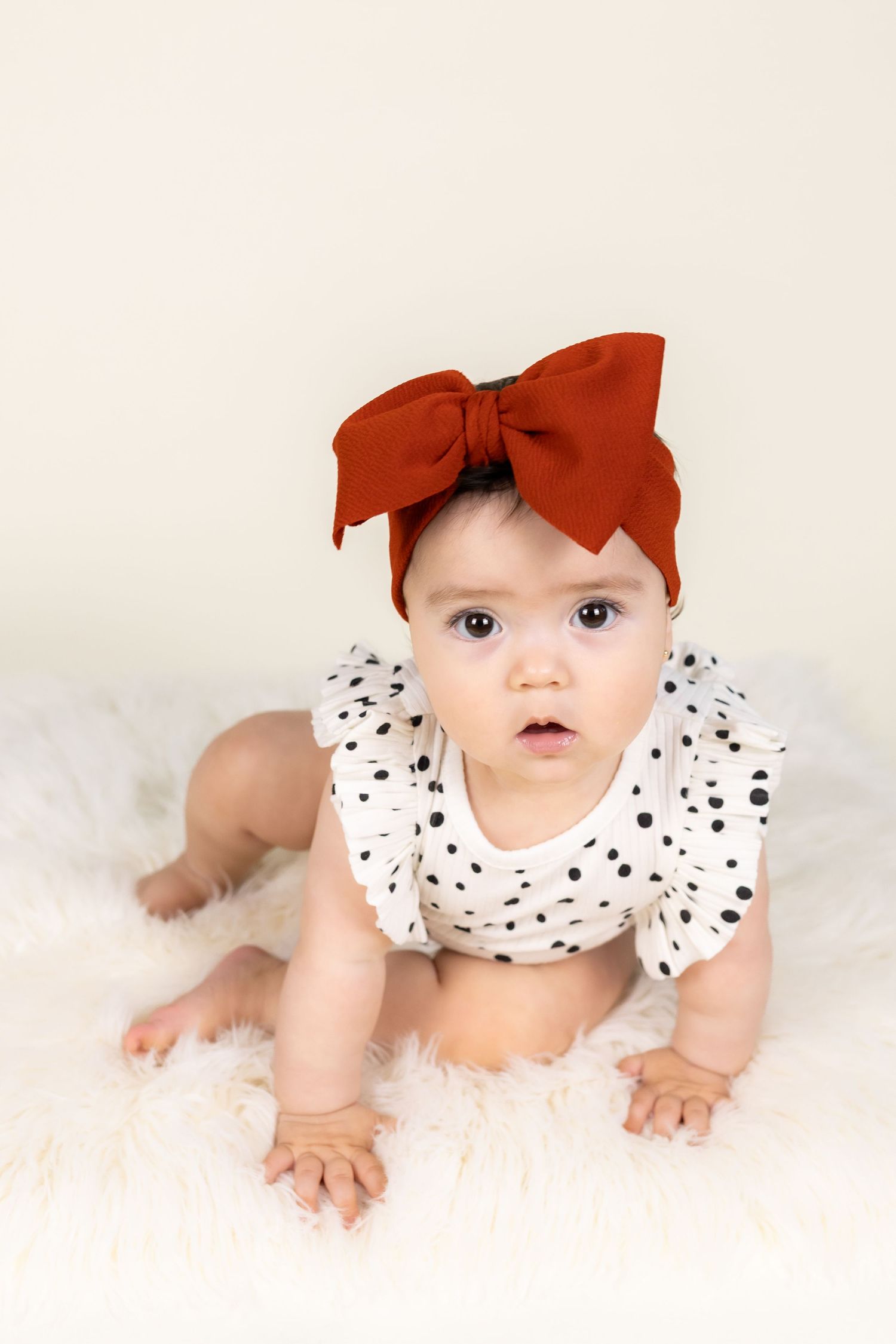 Adorable baby girl in polka dot romper and rust red bow headband, sitting on white fluffy rug.