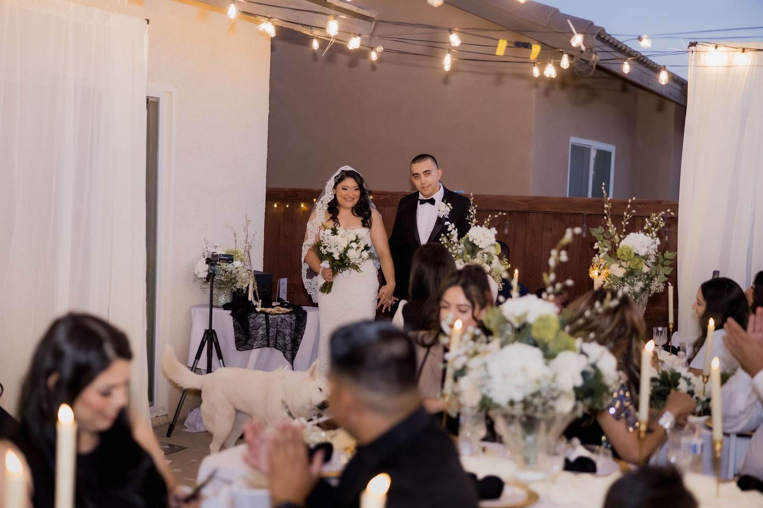 Bride and groom making grand entrance into outdoor wedding reception under string lights.