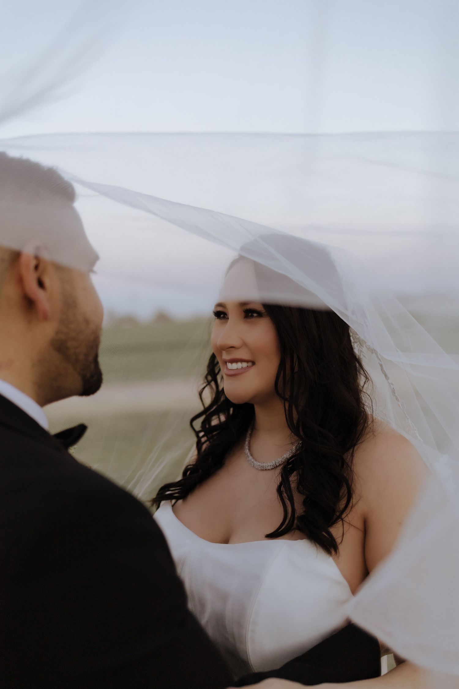 Bride smiles at groom under flowing white veil during romantic outdoor golden hour wedding portrait session.
