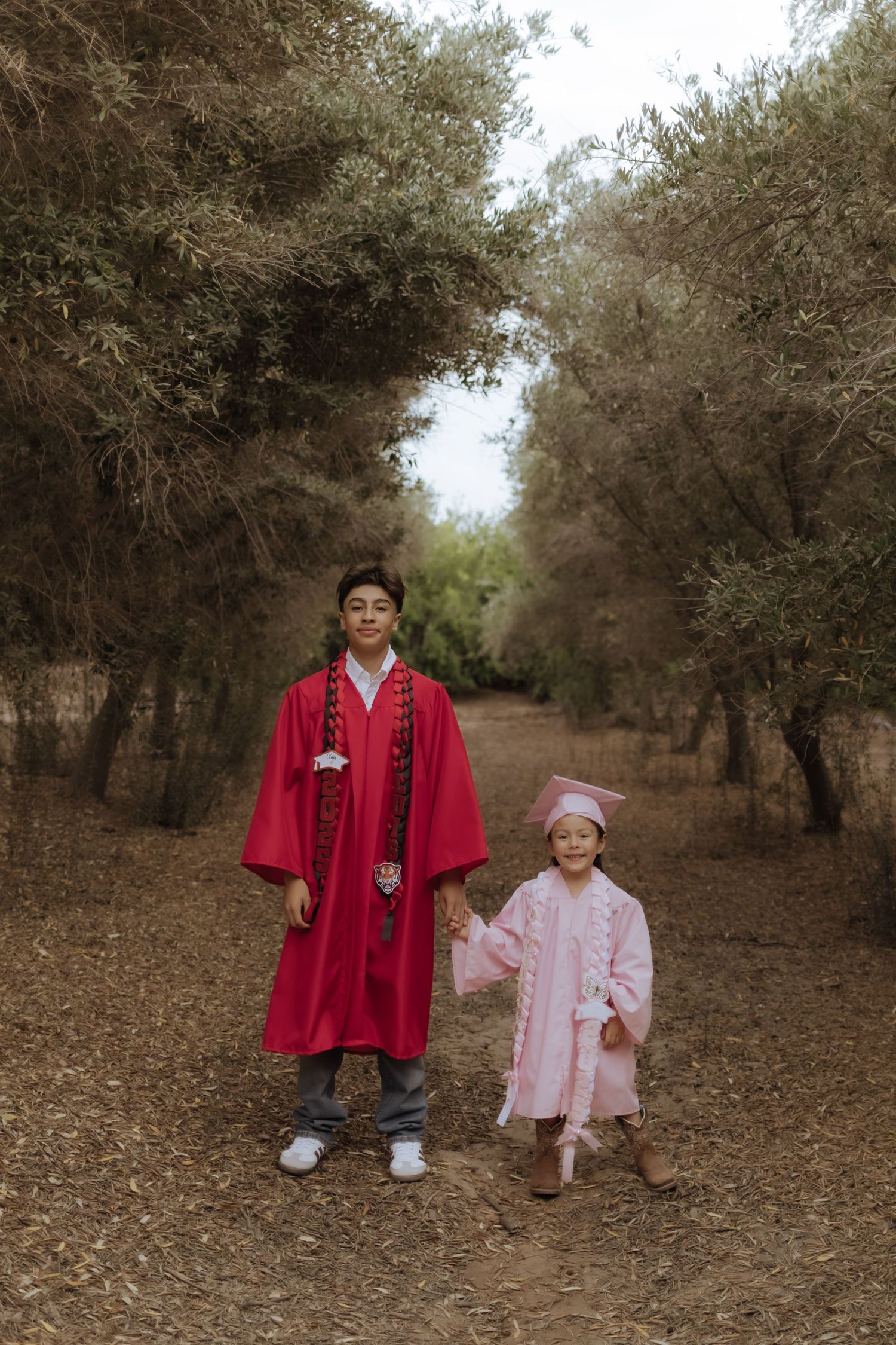 Teen in red graduation gown holds hands with young girl in pink cap and gown on a tree-lined dirt path.