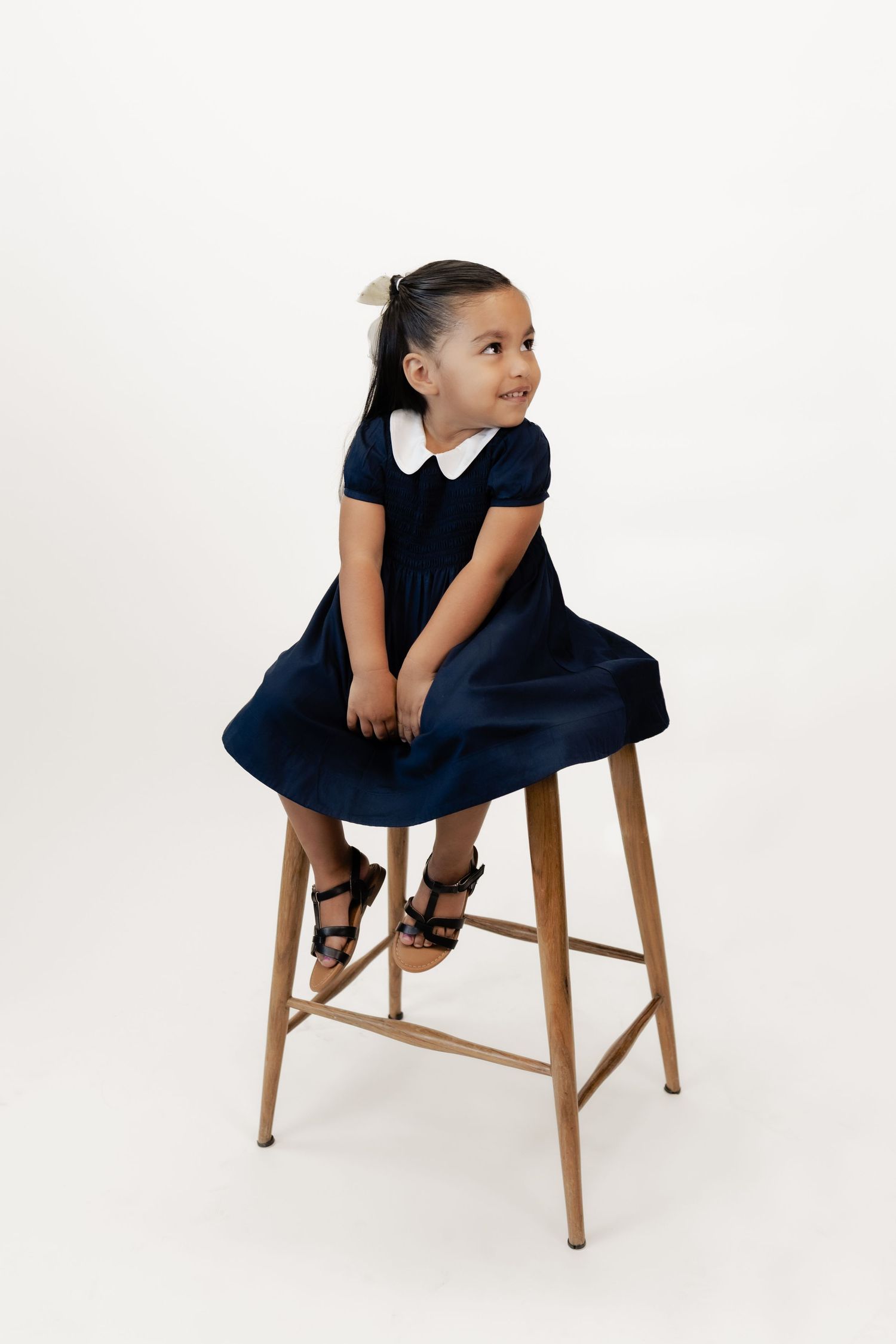 Young girl in navy dress with white collar sits on wooden stool, looking sideways, white background.