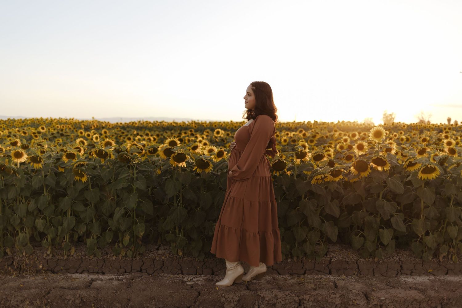 Woman in rust brown dress standing beside blooming sunflower field at golden sunset.