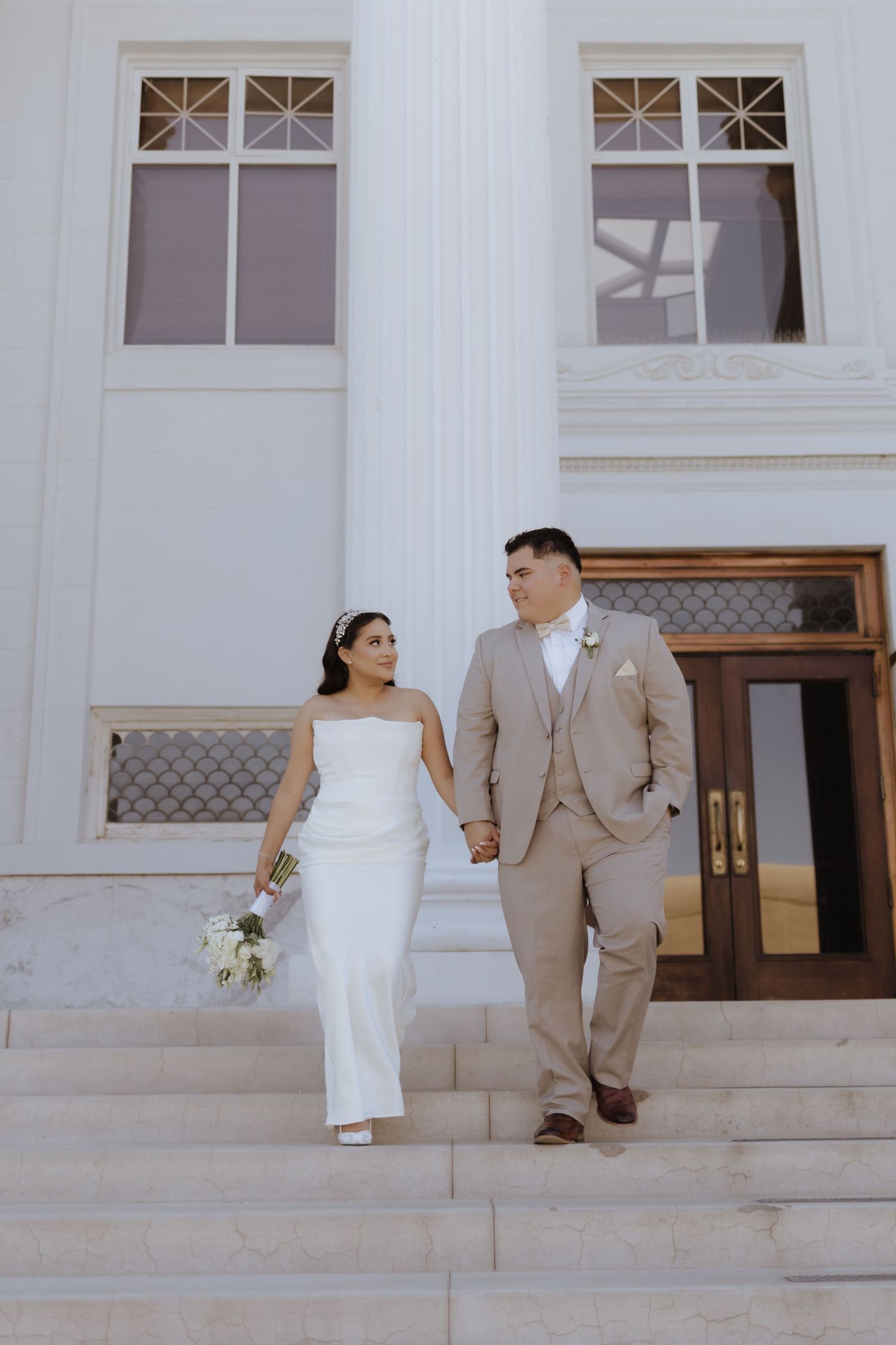 Bride in sleek white gown and groom in tan suit walk hand in hand down steps of white classical building.