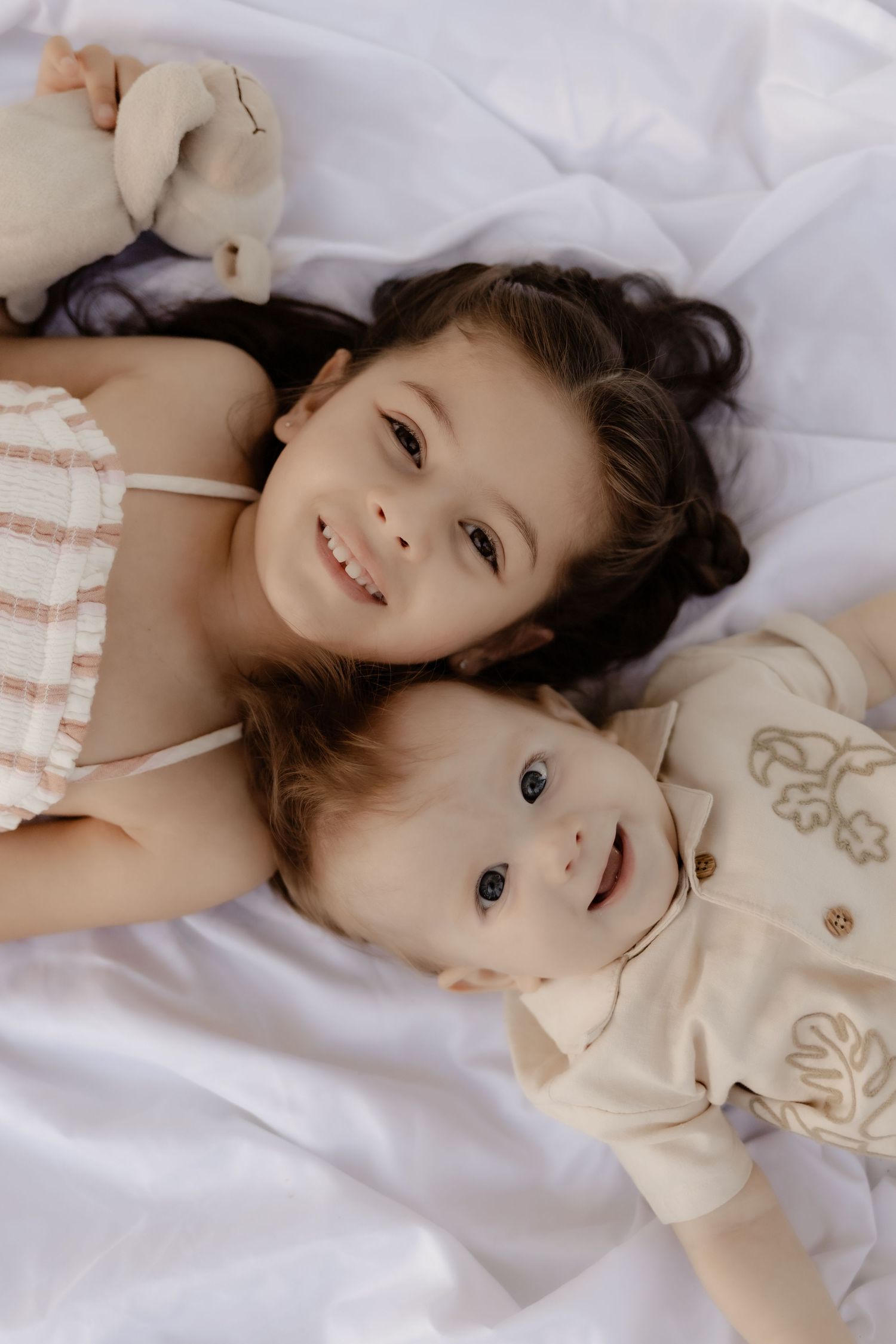 A smiling young girl lies next to her baby sibling on white bedding, both looking up at the camera.