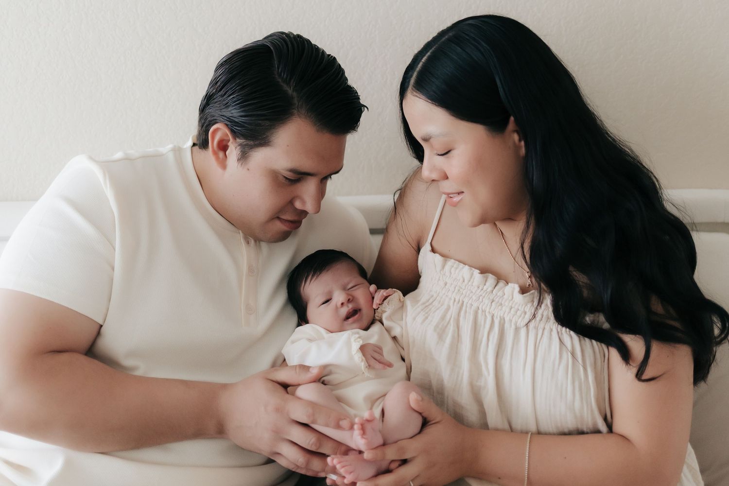 New parents in cream outfits lovingly holding their sleeping newborn baby together.
