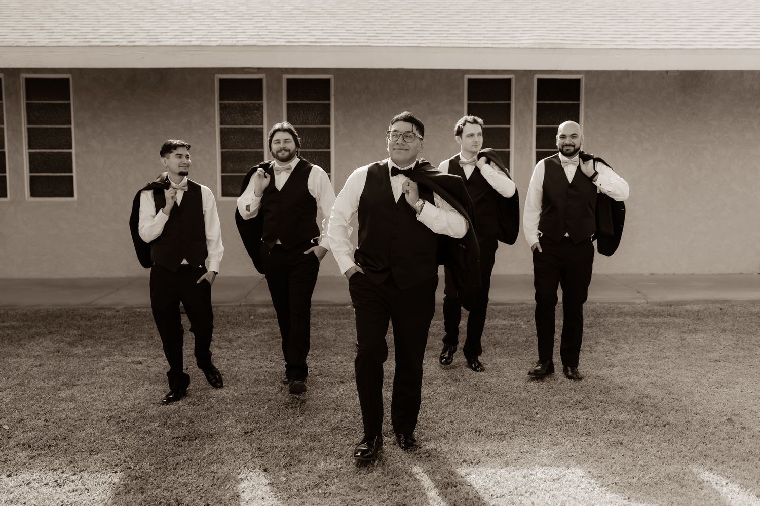 Five groomsmen in black vests and white shirts pose candidly outside a building in a black and white wedding photo.