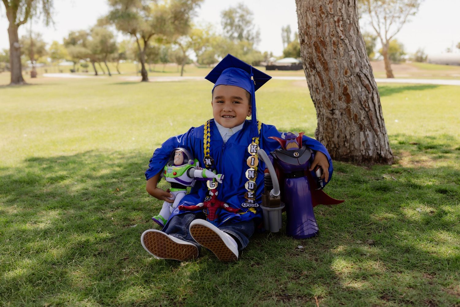 Smiling child in blue graduation cap and gown sits on grass holding Buzz Lightyear toy at kindergarten graduation.