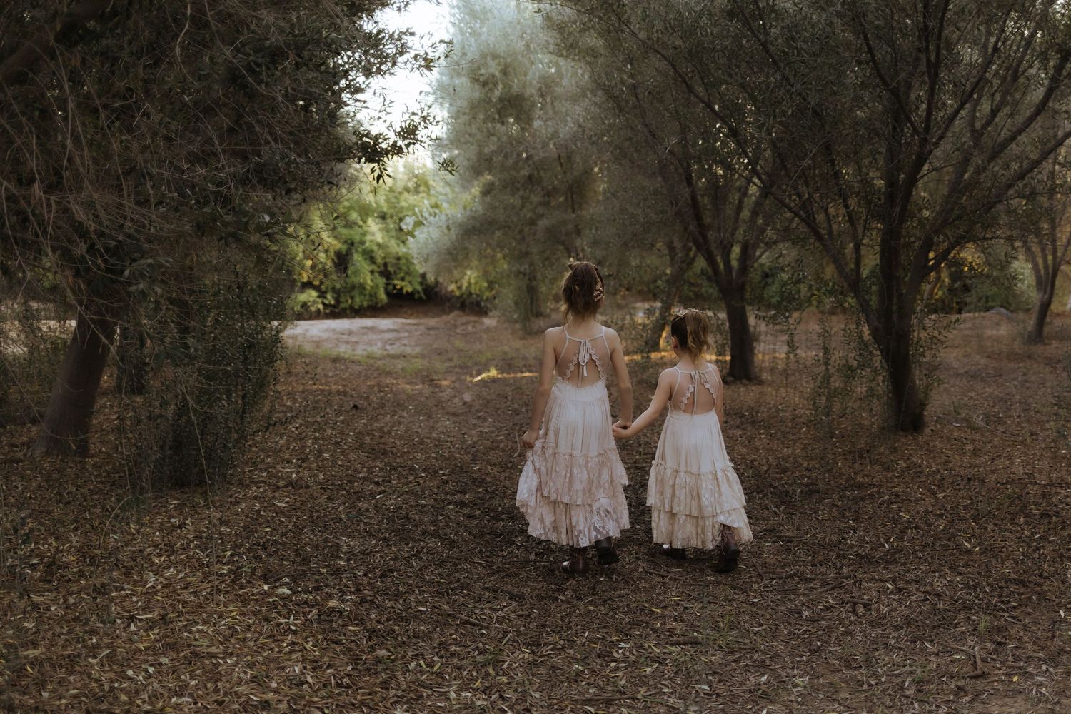 Two young sisters in white lace dresses holding hands walking through wooded forest path.