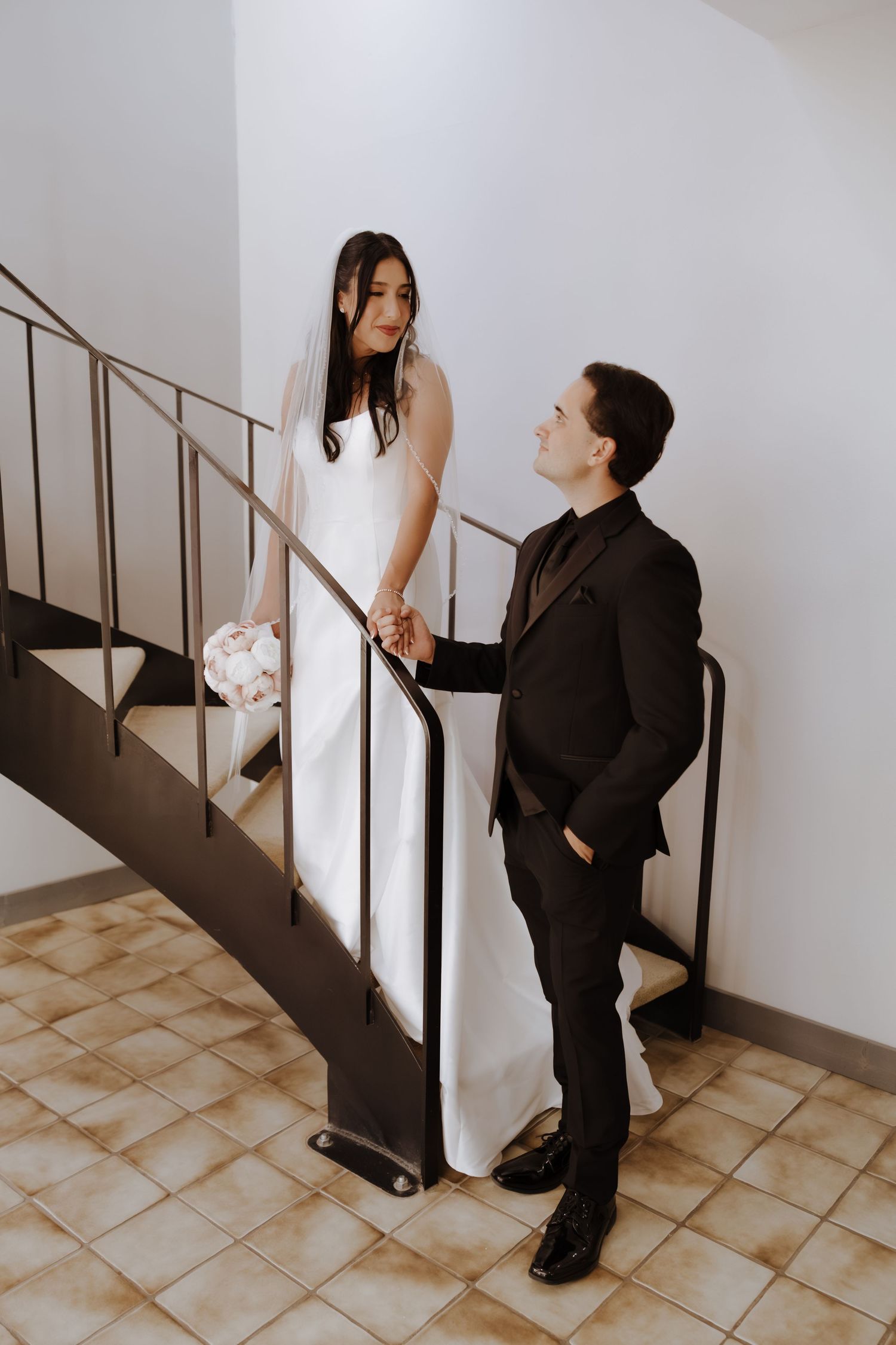 Bride and groom holding hands on a staircase, she in a white gown with veil, he in a black suit.