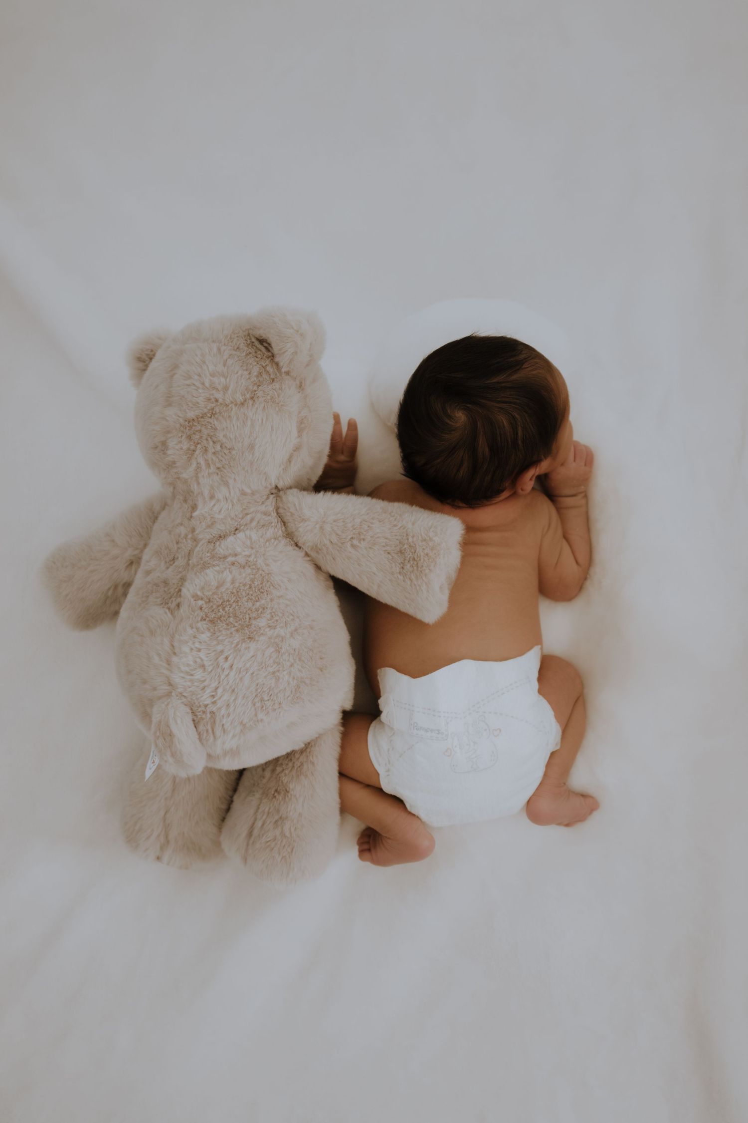 Newborn baby in white diaper sleeping beside a fluffy beige teddy bear on a soft white blanket.