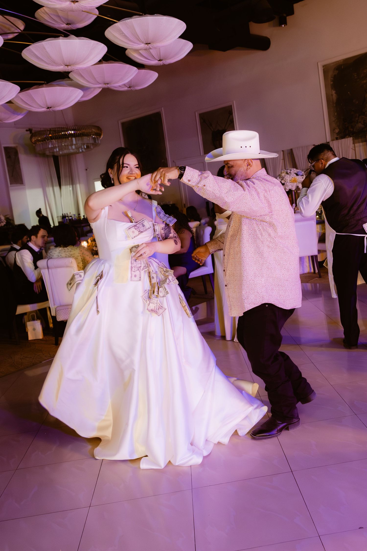 Bride dances with man in white cowboy hat and guayabera during lively wedding reception celebration.