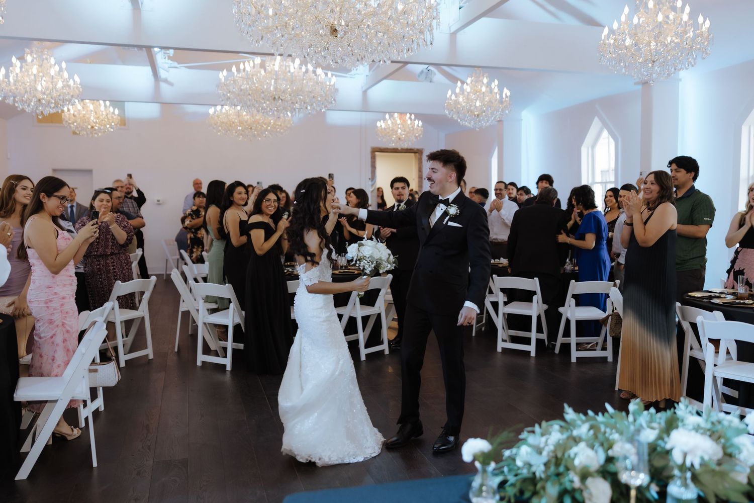 Bride and groom sharing first dance surrounded by cheering guests in chandelier-lit white wedding venue.