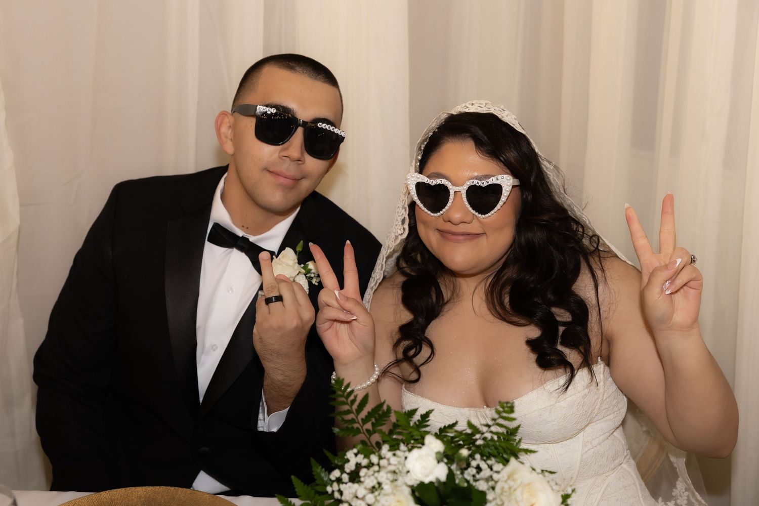 Bride and groom posing playfully with sunglasses making peace signs at wedding photo booth.