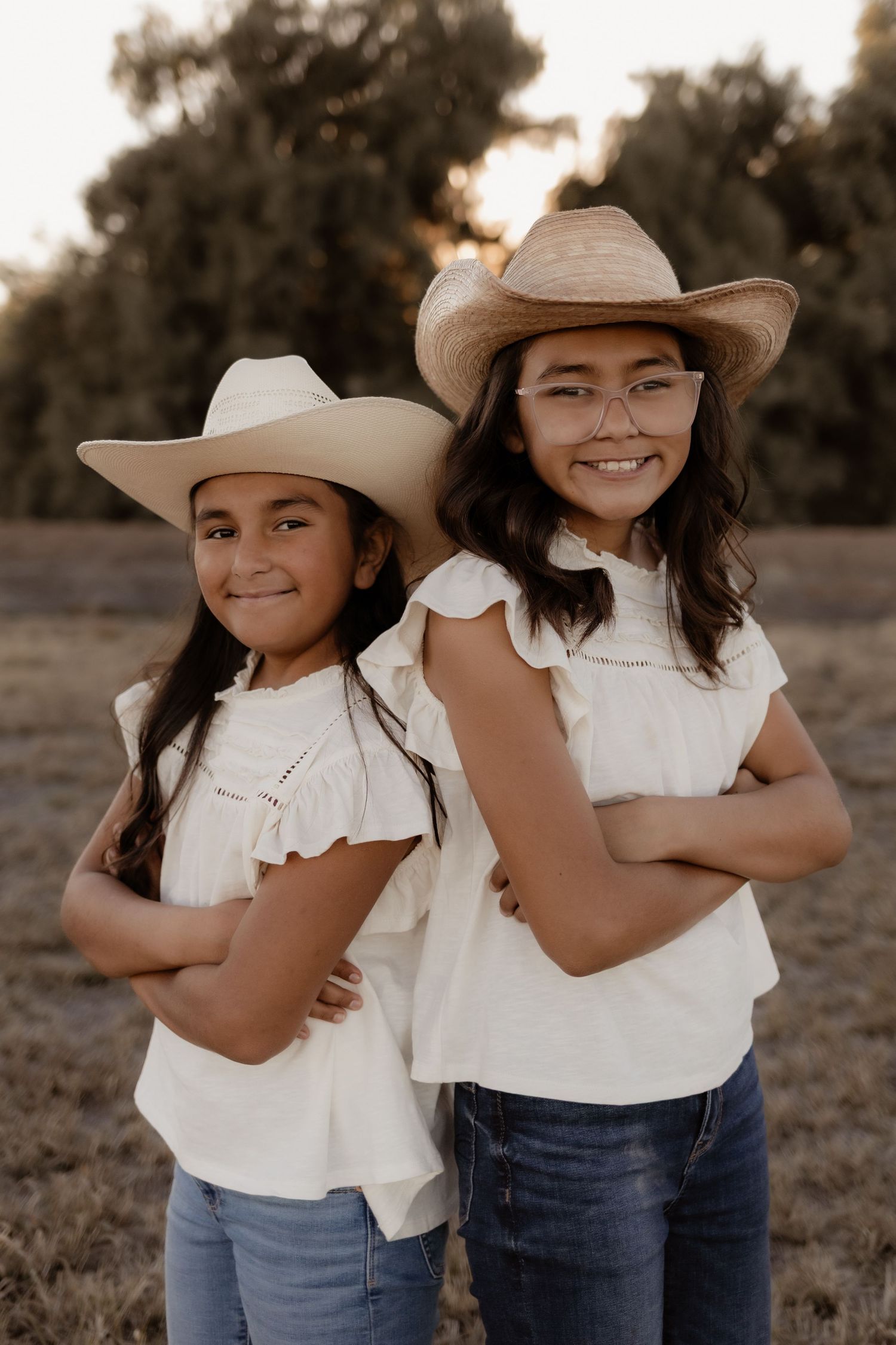 Two girls in white tops and straw cowboy hats posing with arms crossed in a field.