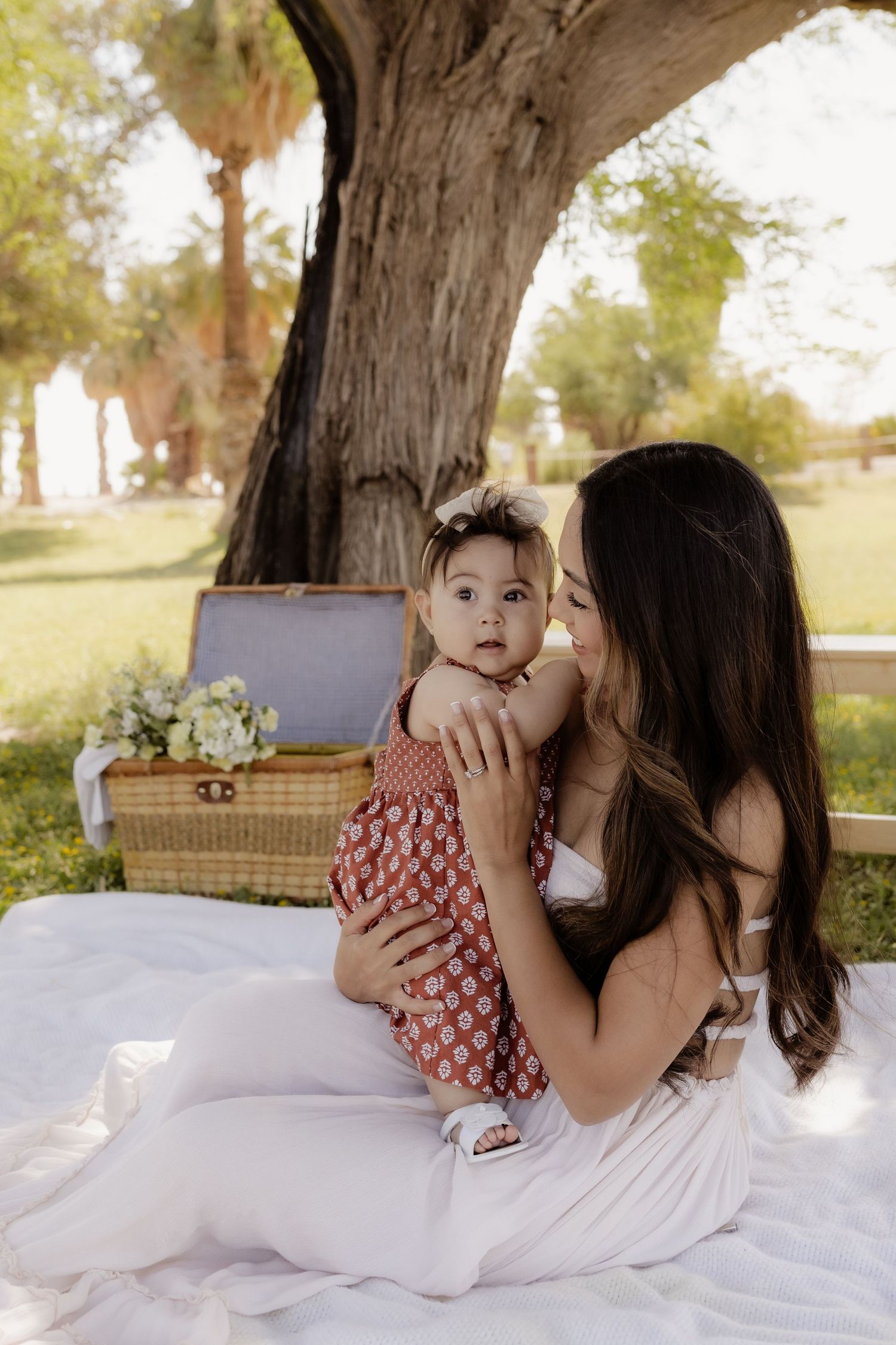 A young mother with long dark hair holds her baby girl while sitting on a blanket under a large tree.