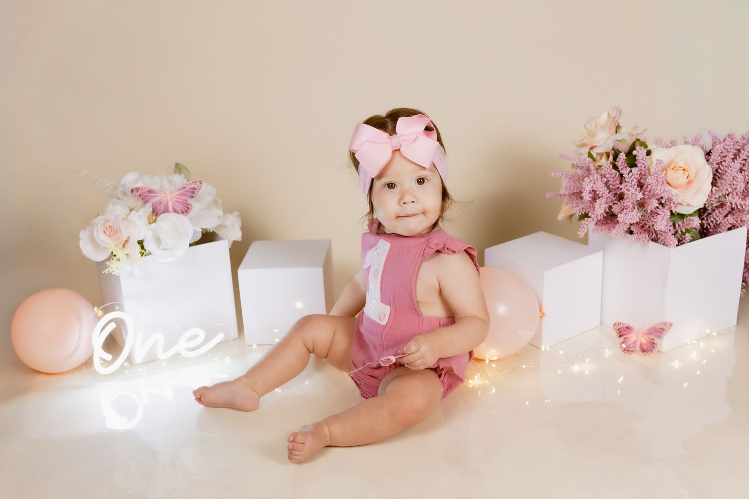 Baby girl in pink romper and bow headband sits among flowers and lights at her first birthday photo shoot.