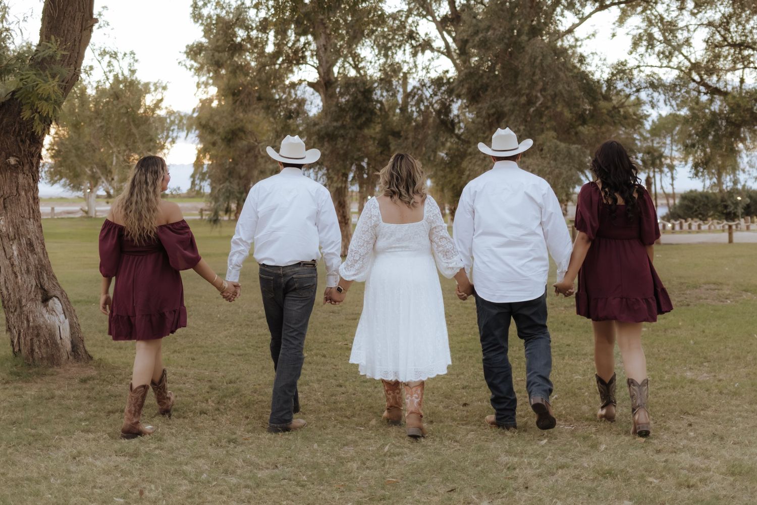 Five people in western attire hold hands walking through a green park from behind.