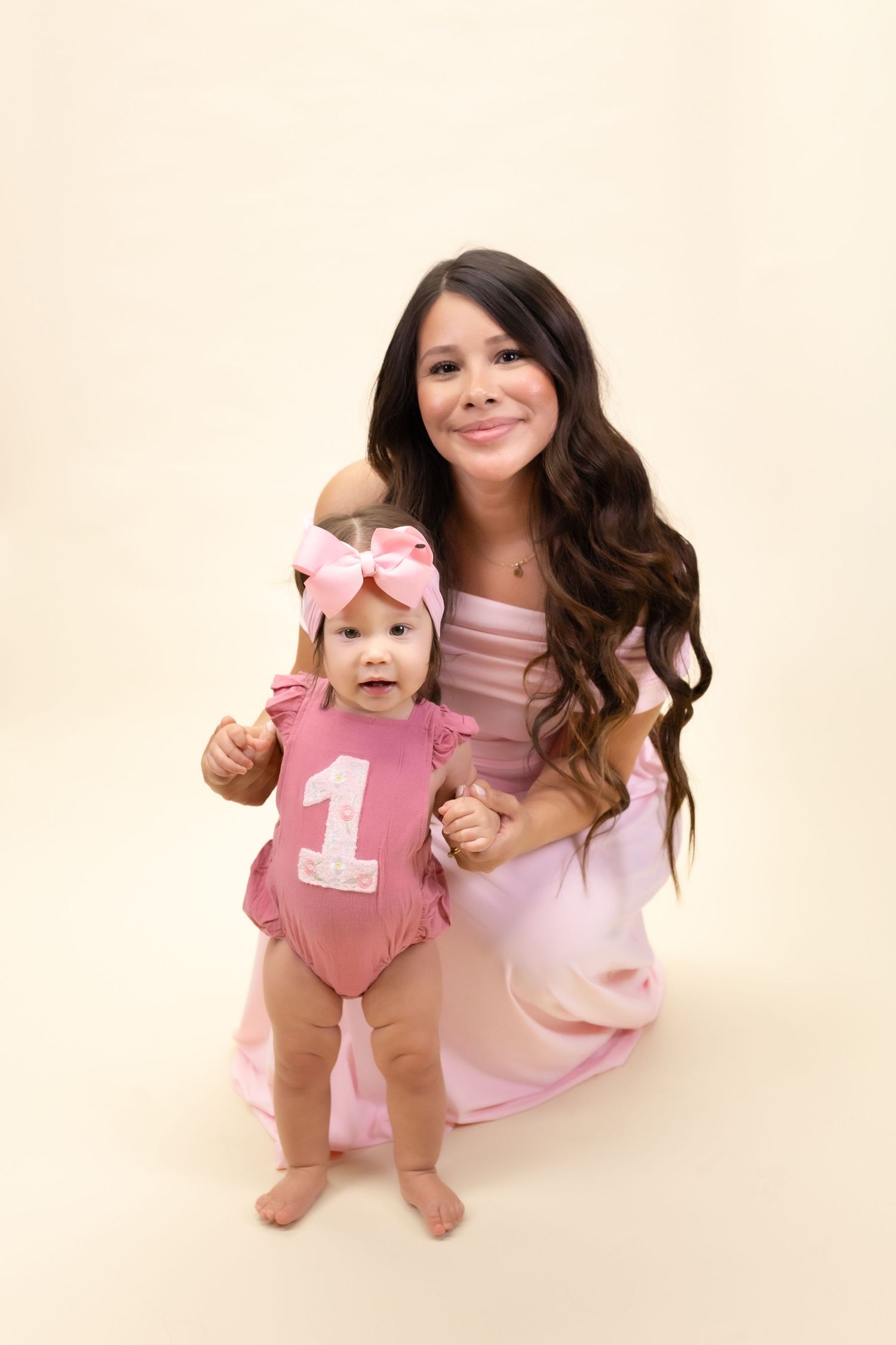 A woman in a pink dress kneels beside a baby in a pink '1' onesie and bow headband for a first birthday photo.