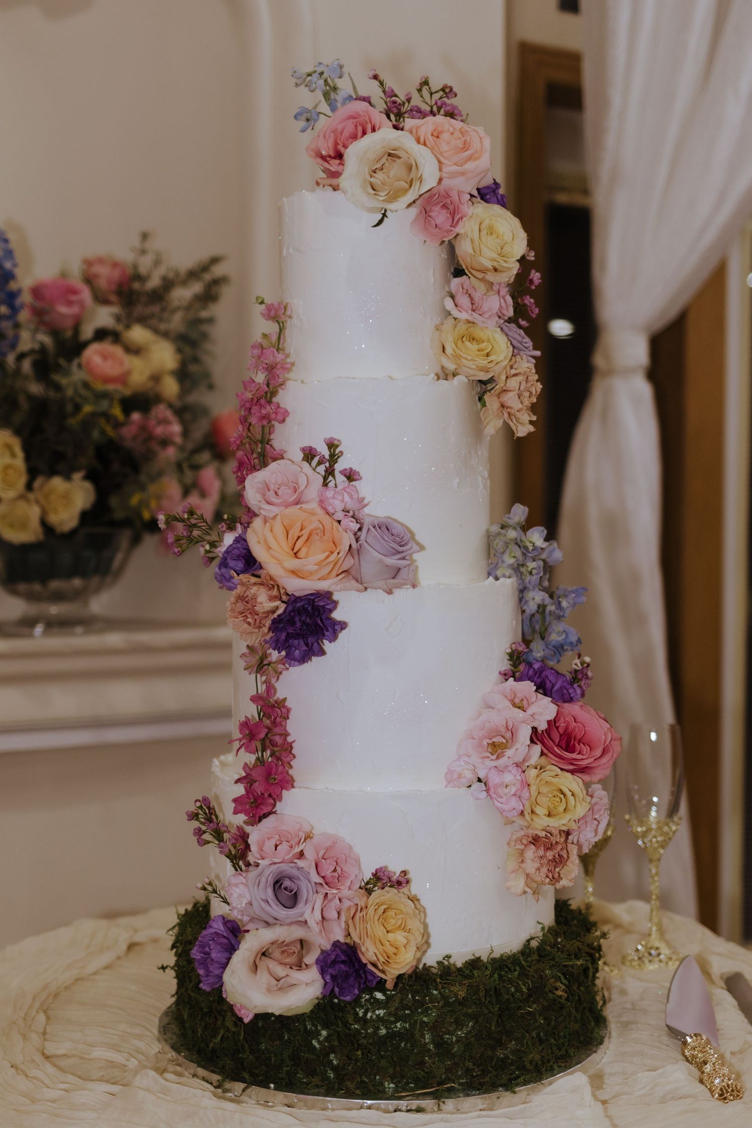 Elegant four-tier white wedding cake decorated with cascading colorful roses and flowers on a moss base.