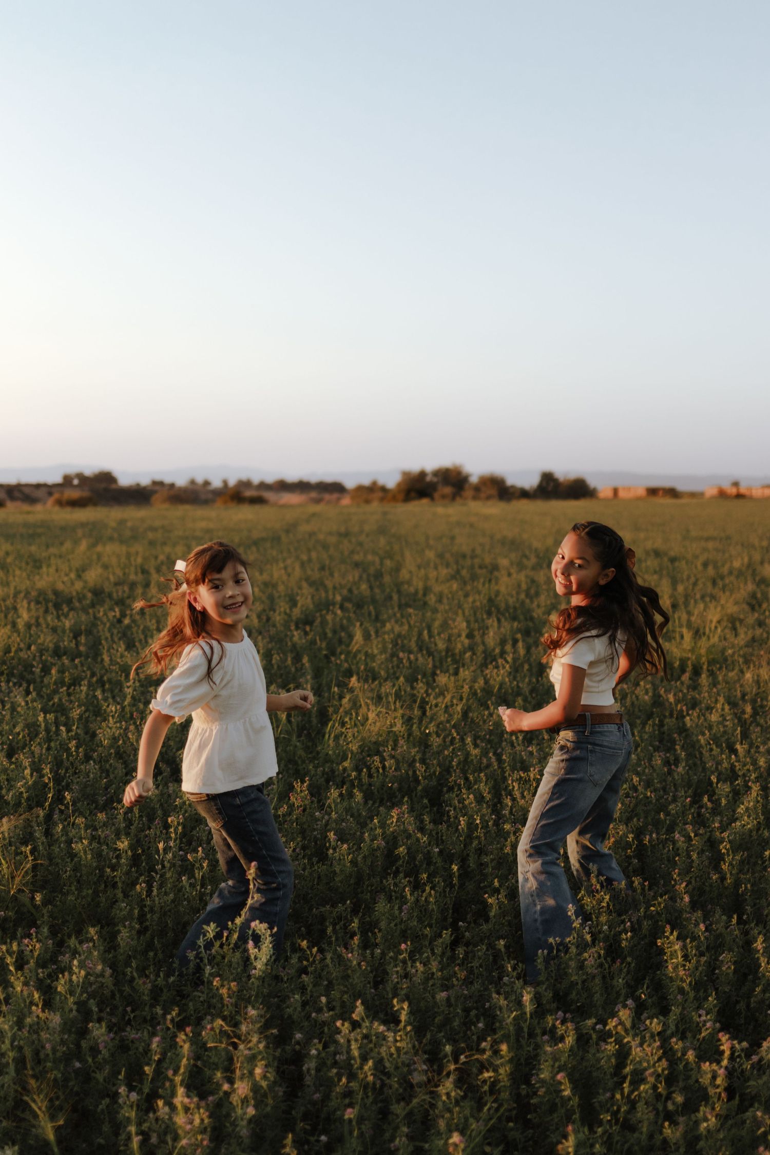 Two girls in white tops and jeans laugh and run through a green field at golden hour sunset.