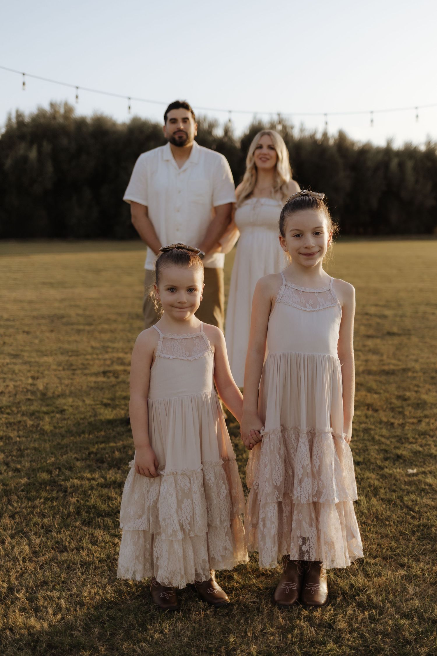 Family of four with two girls in matching cream dresses posing in a golden field at sunset.