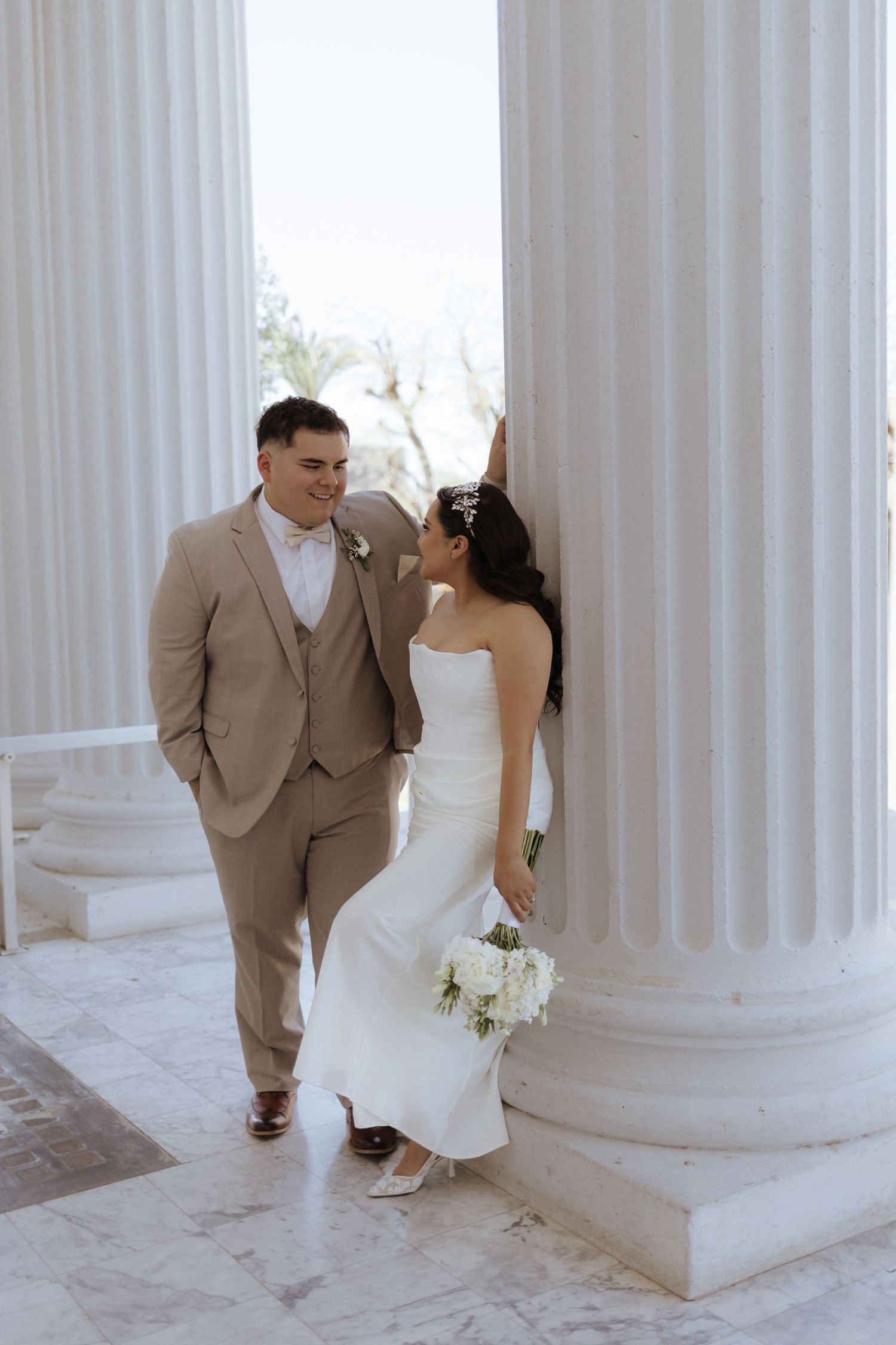 Bride and groom pose between white marble columns, she in a strapless gown holding white roses, he in a tan suit.