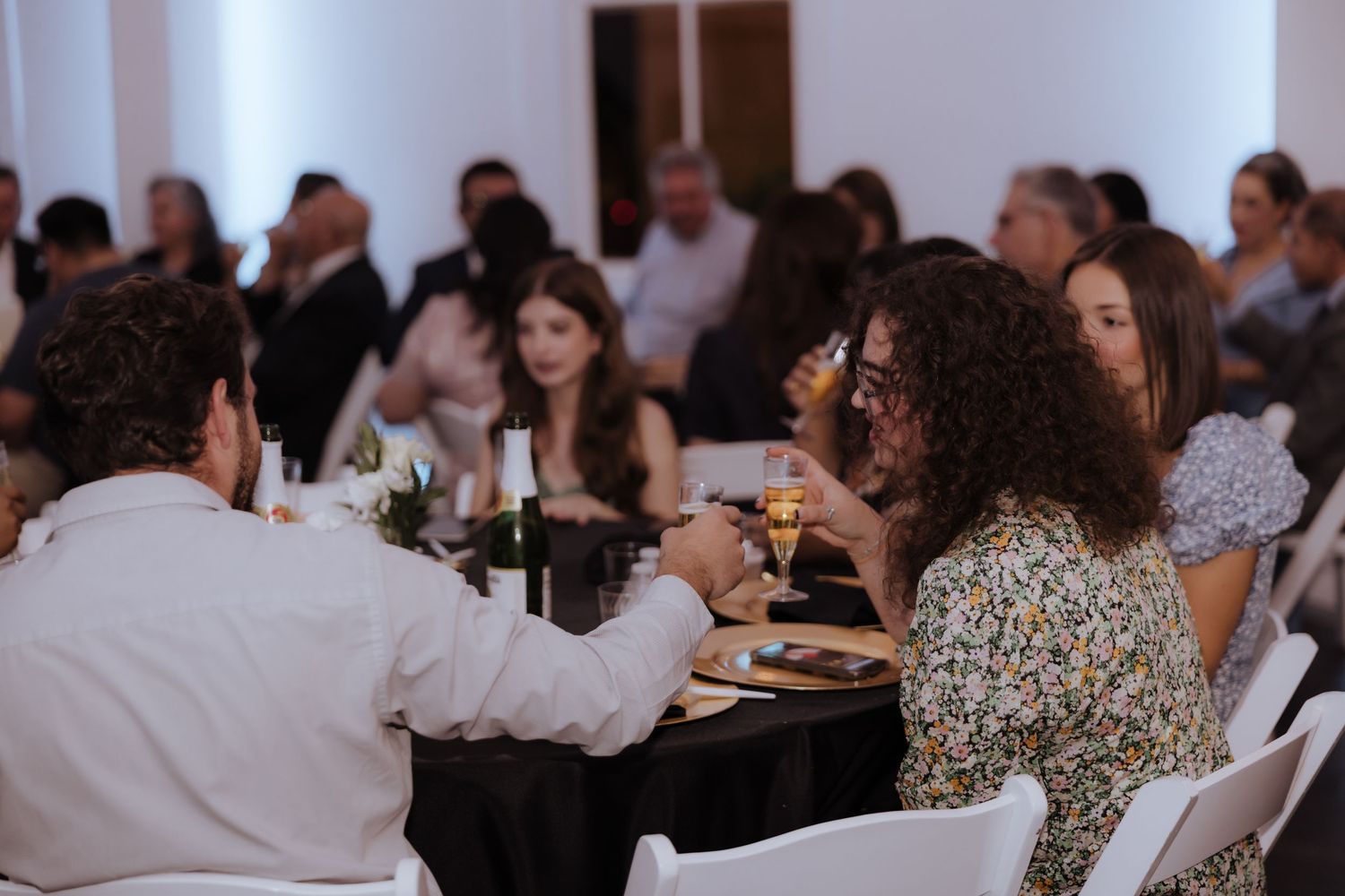 Wedding reception guests seated at black-draped tables, enjoying drinks and conversation in elegant venue.