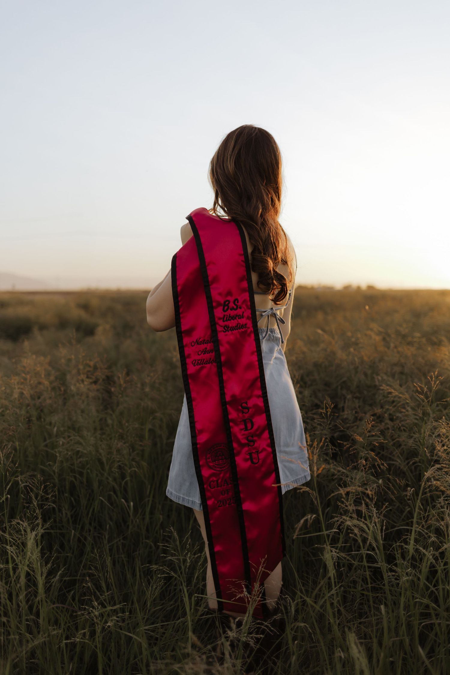 Graduate holds red satin stole while standing in golden field at sunset, viewed from behind.