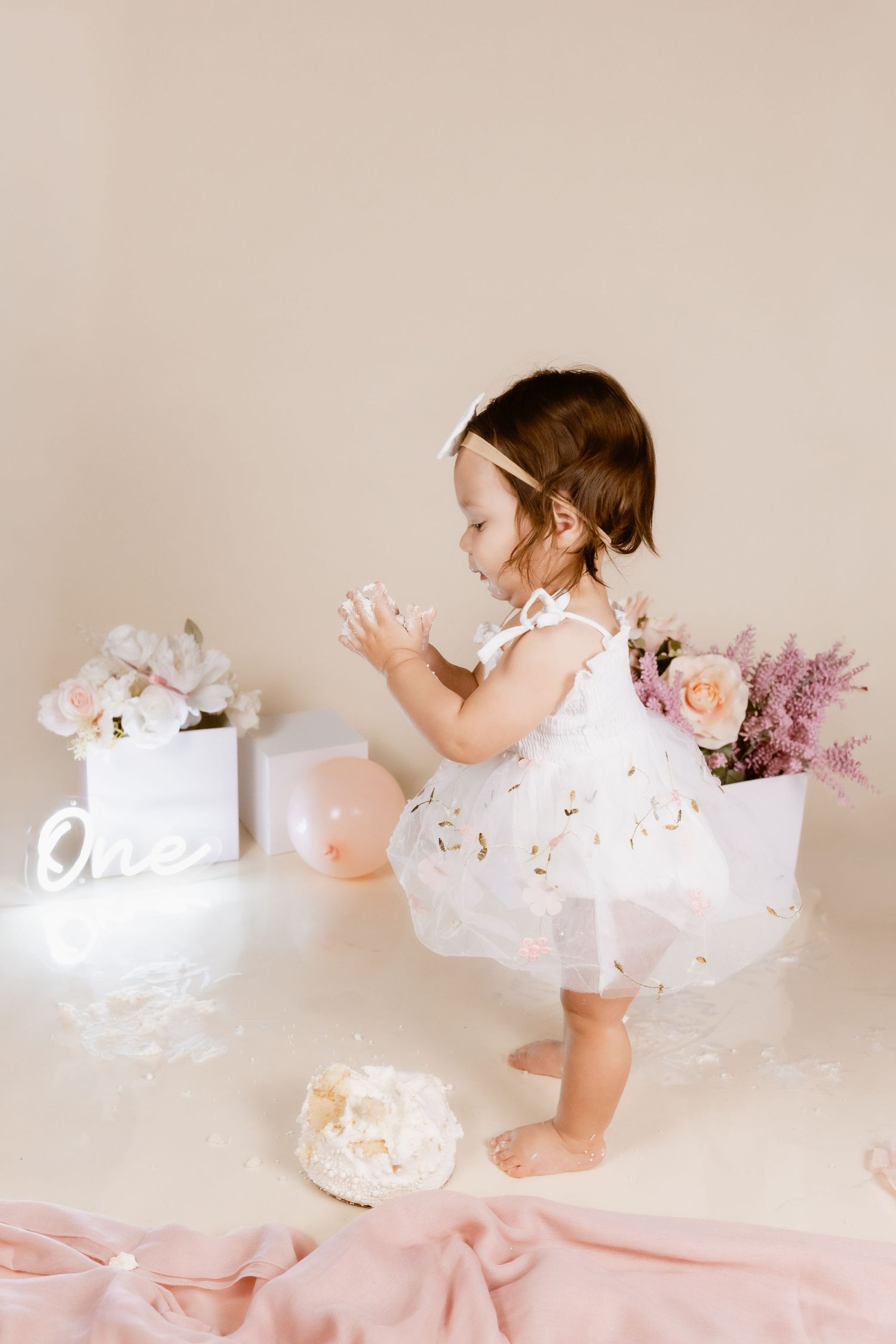 Toddler girl in white dress clapping at first birthday cake smash with floral decorations and One sign.