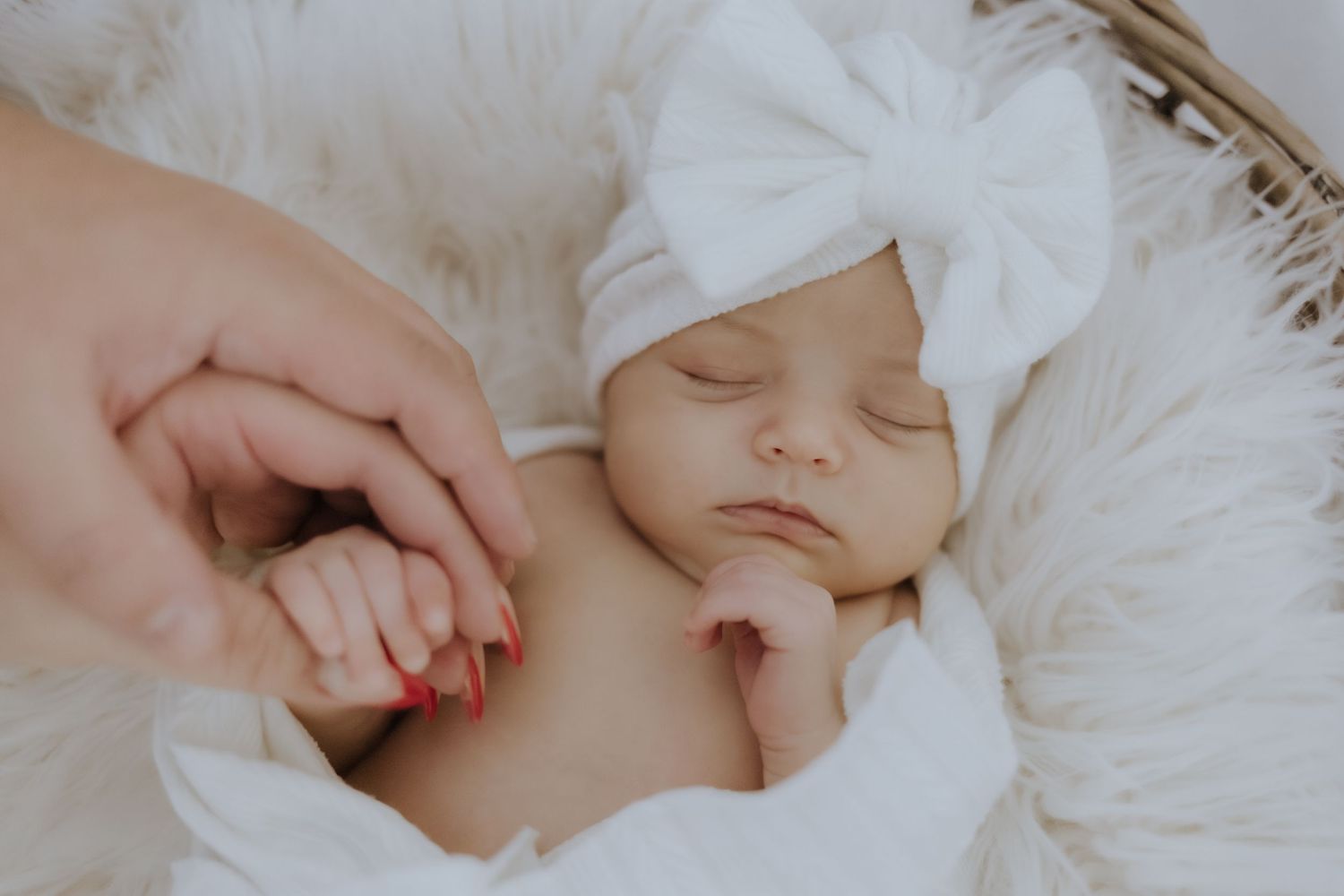 Sleeping newborn baby in white bow headband held in a soft basket with mother's hand.