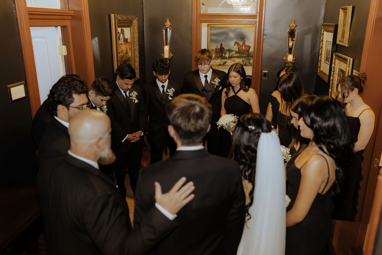 Wedding party gathered in a circle for a group prayer in an elegantly decorated indoor venue.