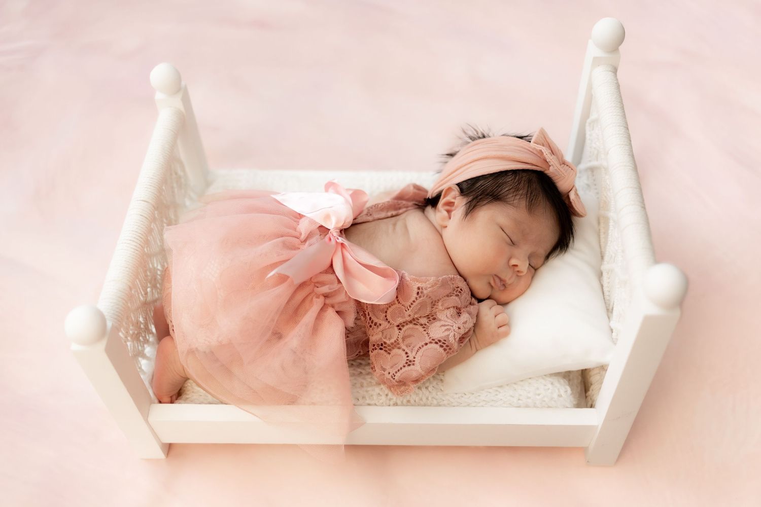 Newborn baby girl sleeping peacefully in a white mini bed, wearing a pink tutu dress and bow headband.