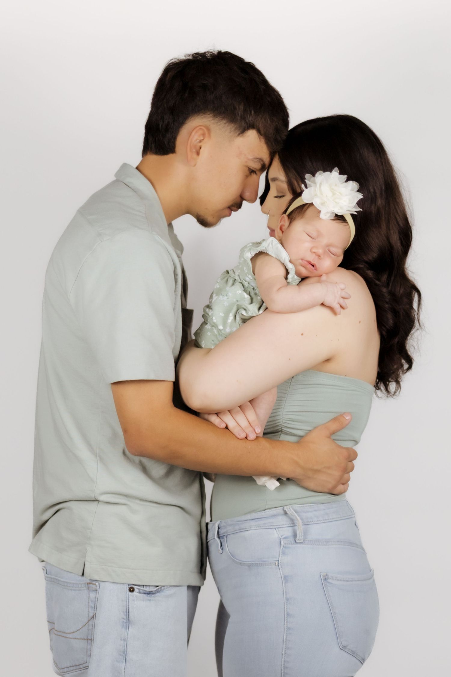 A young couple in sage green tops tenderly hold their sleeping newborn baby girl with a white flower headband.