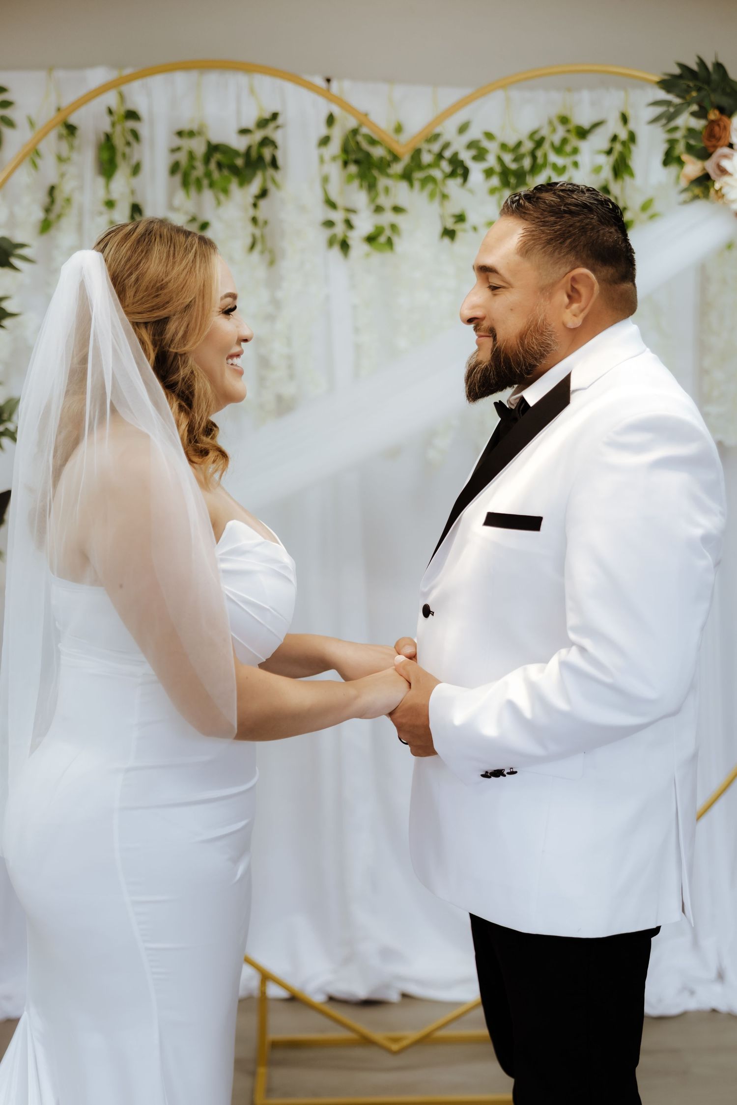 Smiling bride and groom holding hands at altar beneath heart-shaped floral arch during ceremony.