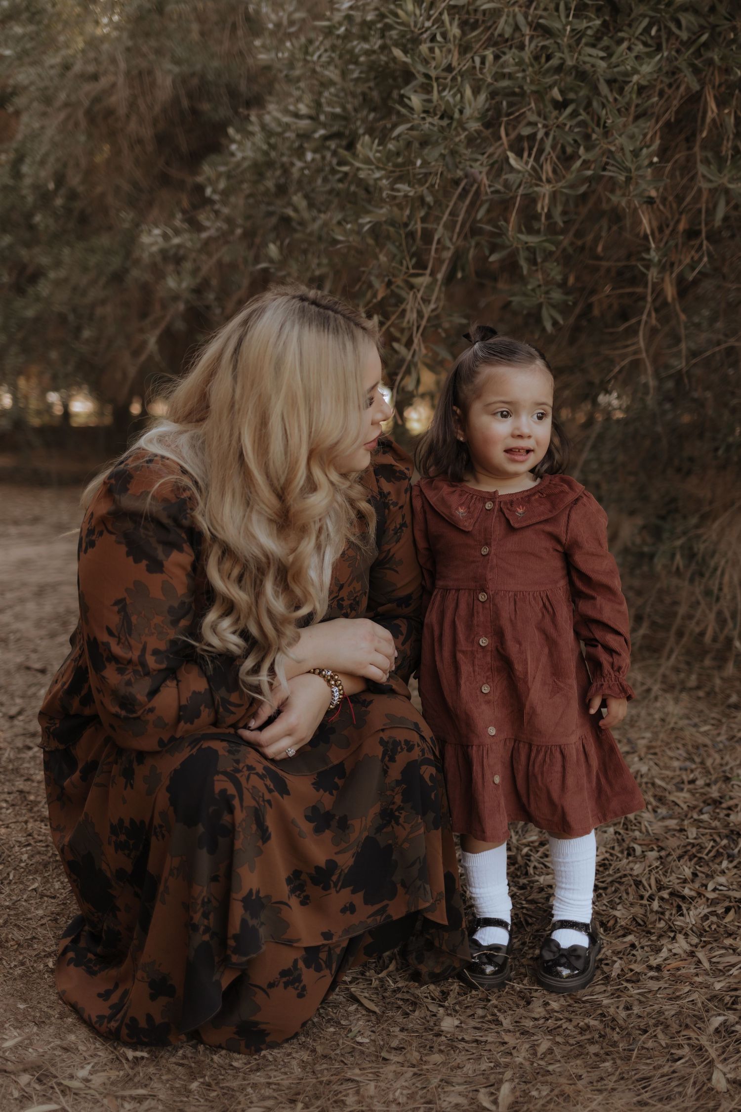 Blonde mother in floral brown dress crouching beside toddler girl in rust dress under trees.