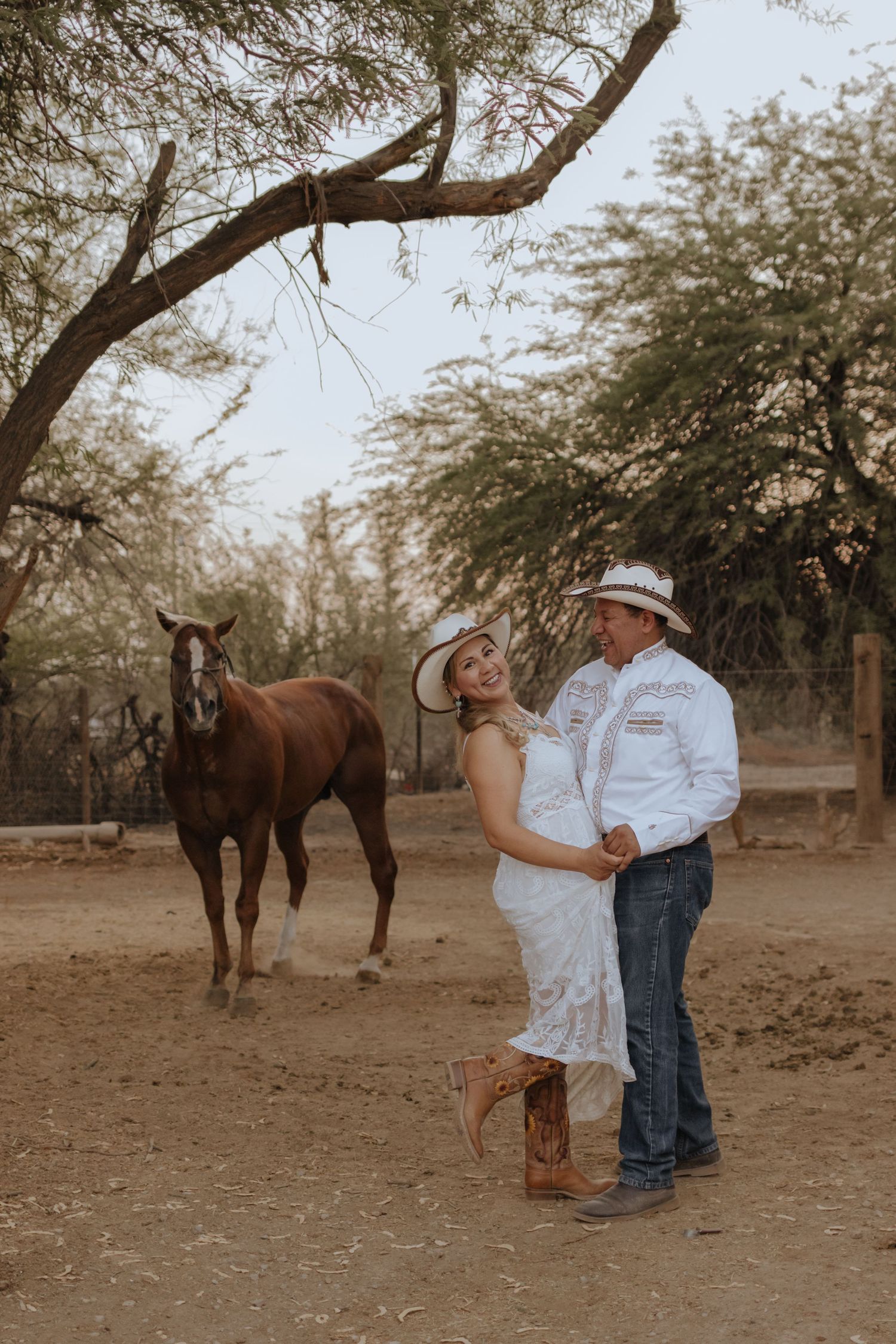 Couple in cowboy hats dancing near a chestnut horse under a tree in a rustic outdoor setting.