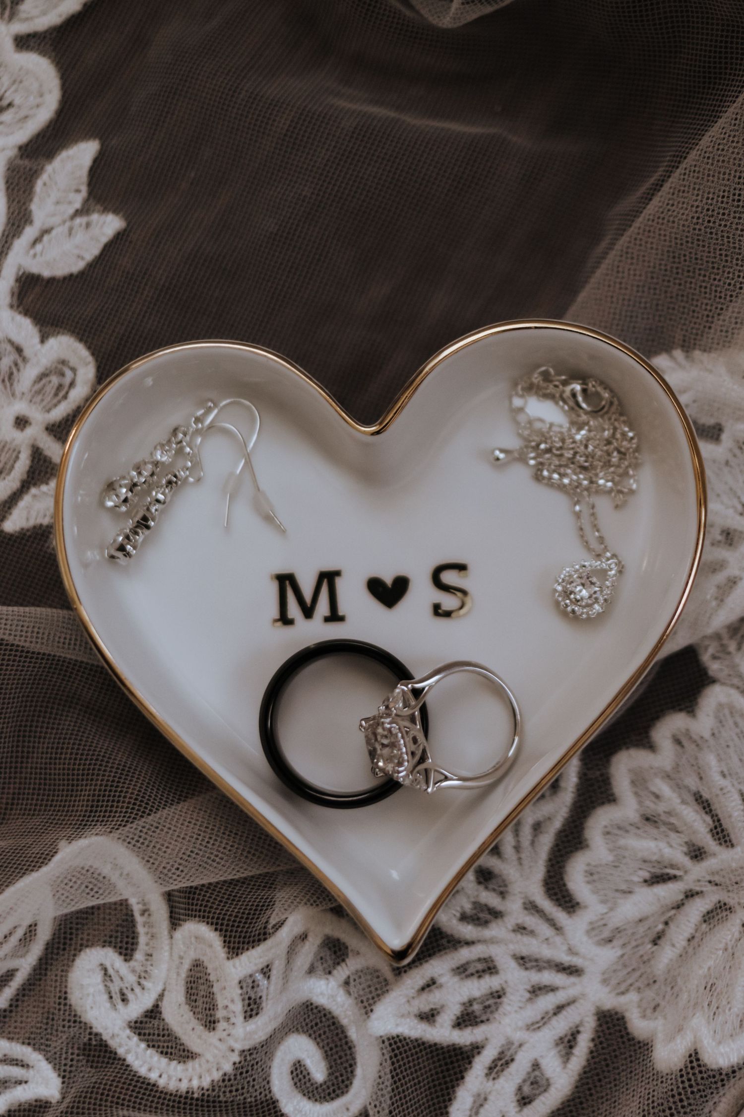 Heart-shaped ring dish monogrammed with M and S holds wedding bands, diamond ring, and silver earrings on lace veil.