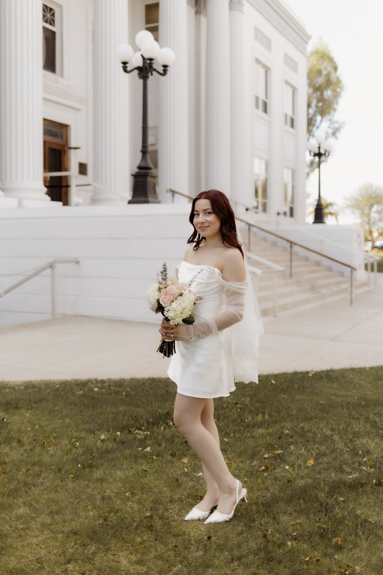 Bride in short white off-shoulder dress holding floral bouquet standing on grass outside white building.
