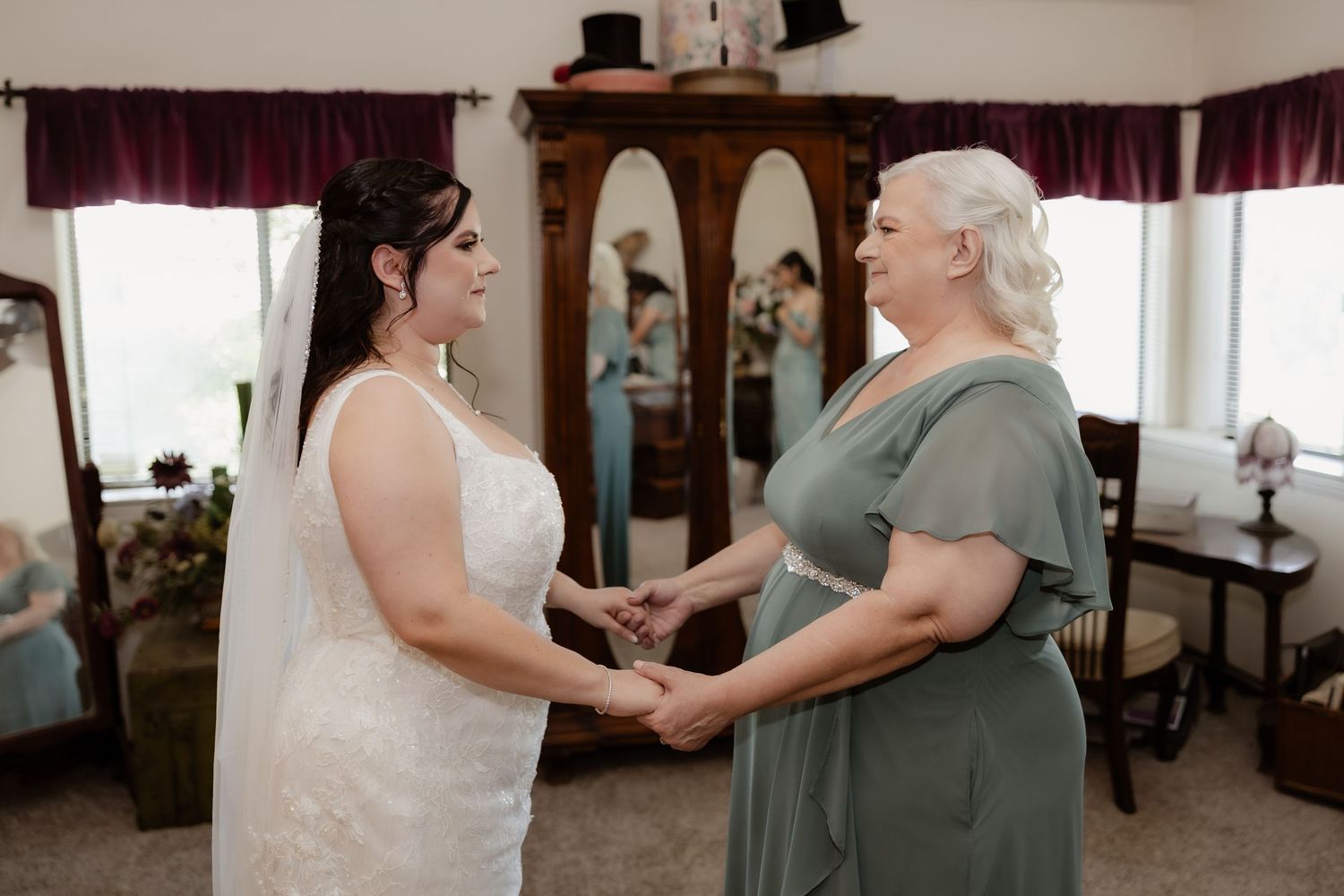 Bride in white lace wedding gown holds hands with a woman in sage green dress during an emotional pre-ceremony moment.