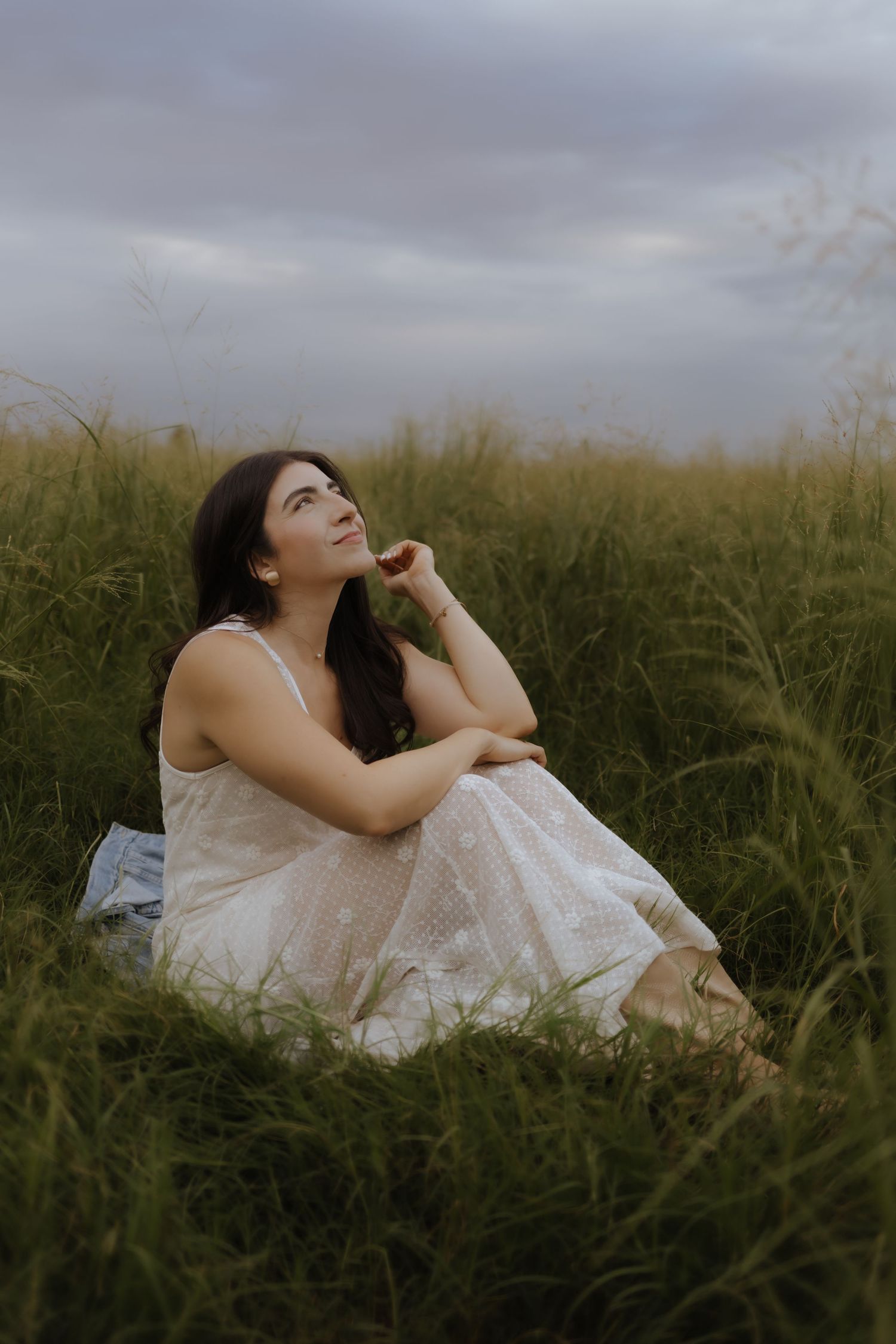 Young woman in white dress sitting in tall green grass looking up at cloudy sky.