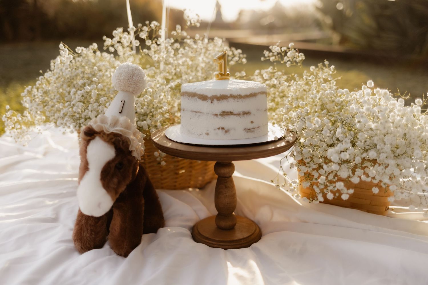 Naked white birthday cake on wooden stand surrounded by baby's breath flowers outdoors.