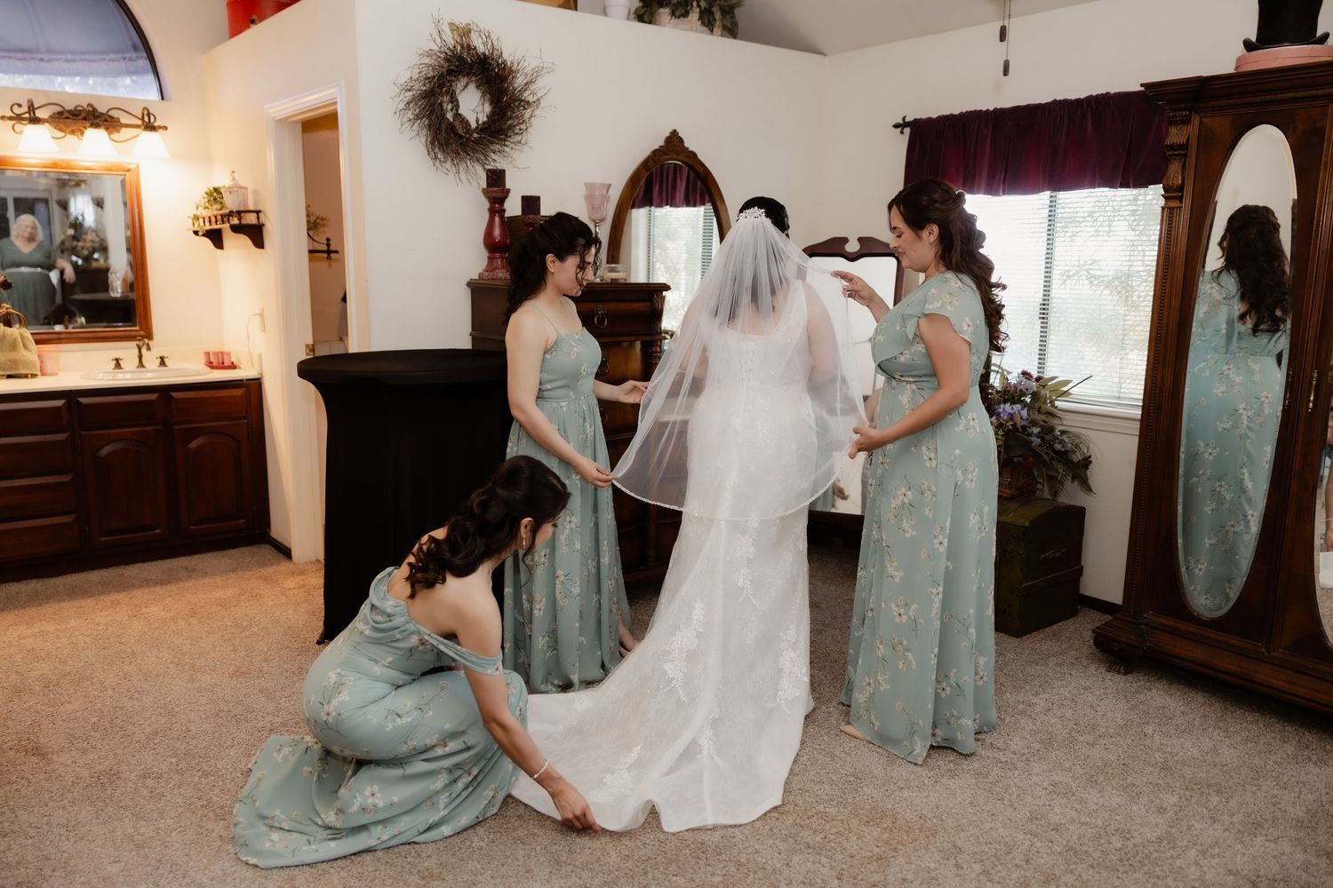 Bridesmaids in sage green dresses help the bride with her veil and dress in a warmly lit getting-ready room.