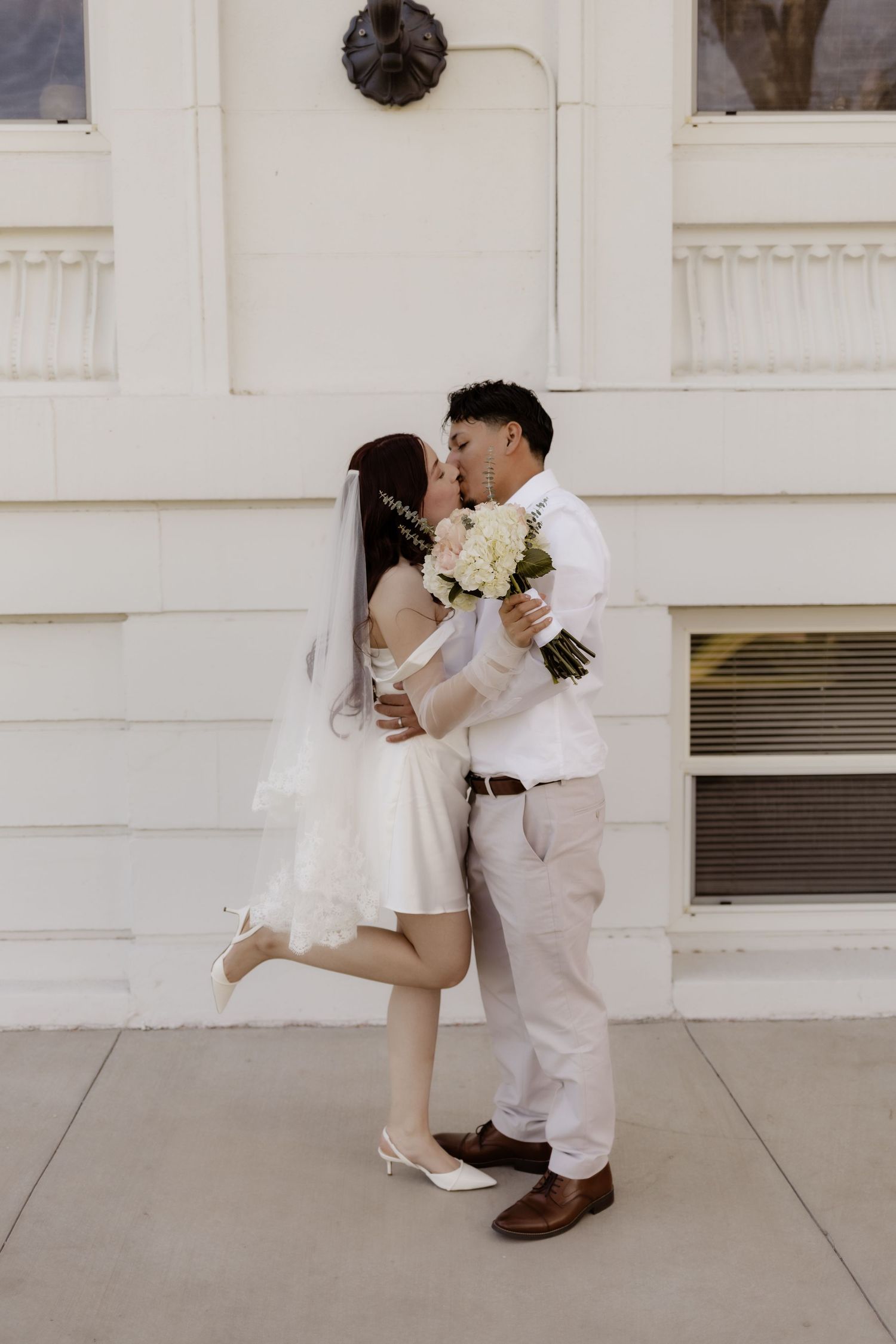 Bride and groom sharing a kiss outside white building, bride in mini dress holding white bouquet.