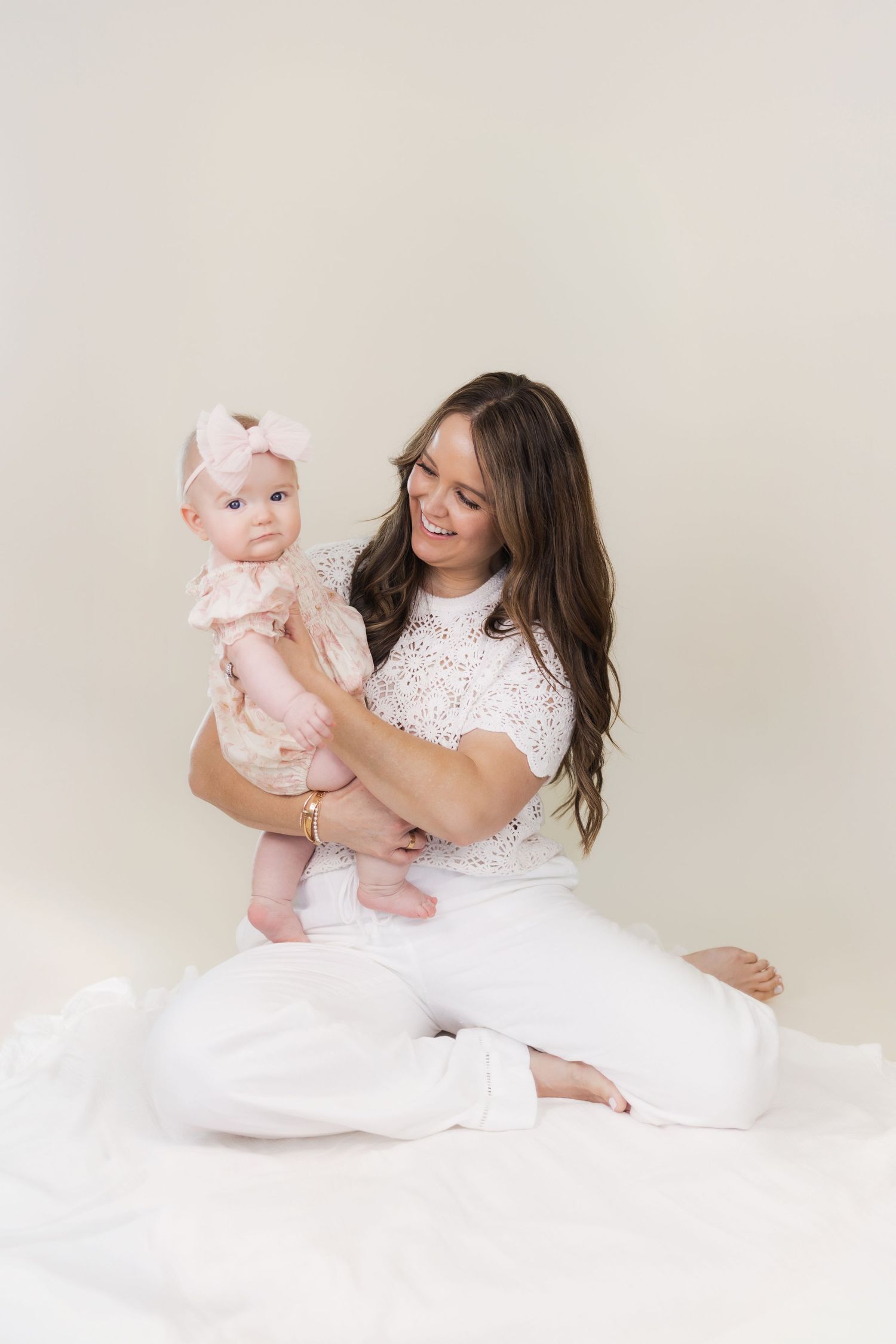Smiling brunette mother in white outfit sitting on floor holding adorable baby in pink dress.