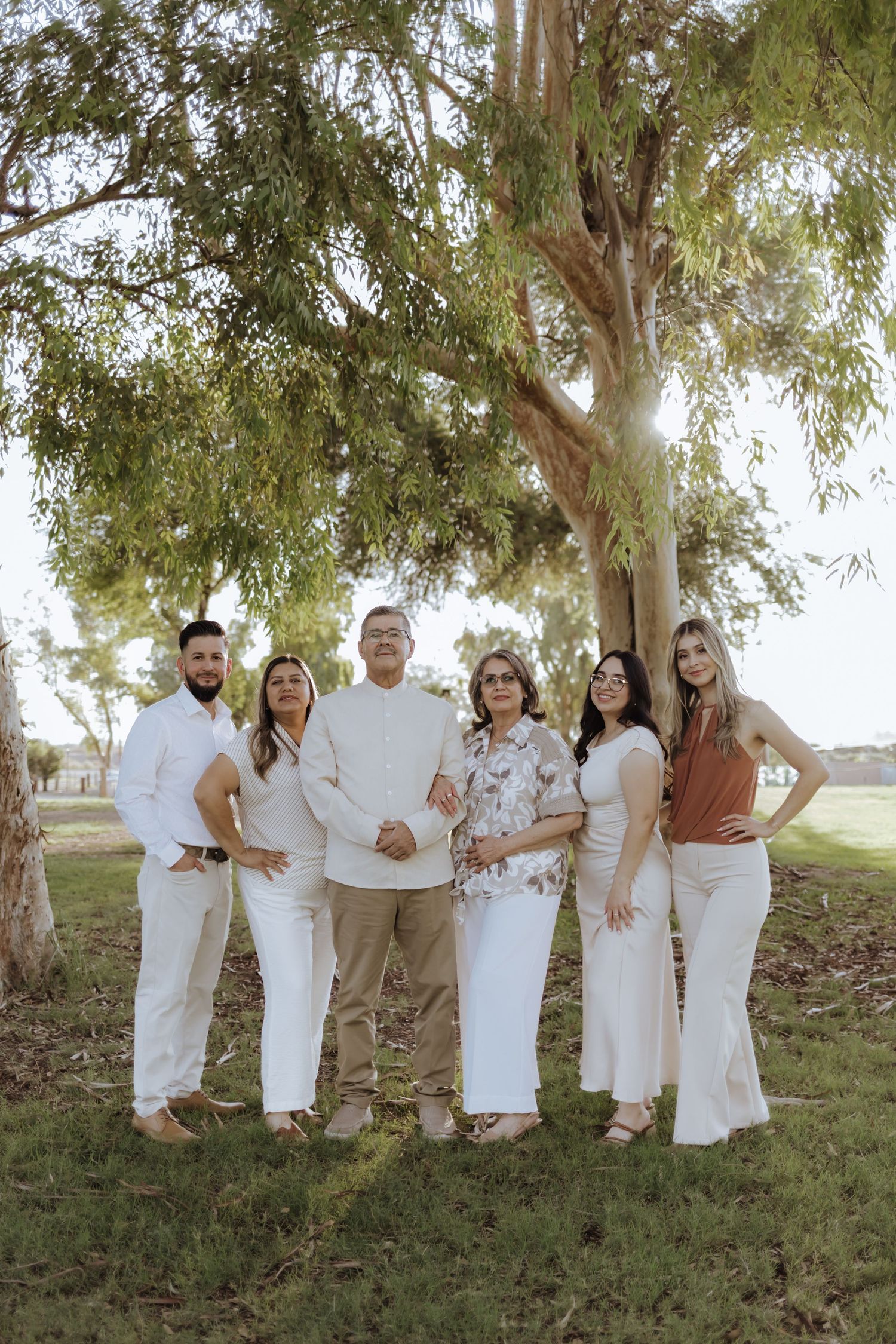 Family of six poses together smiling under large trees in a sunny park setting.