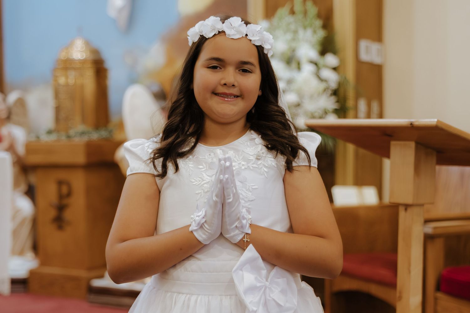 Young girl in white dress and floral crown prays with hands together at First Communion ceremony in church.