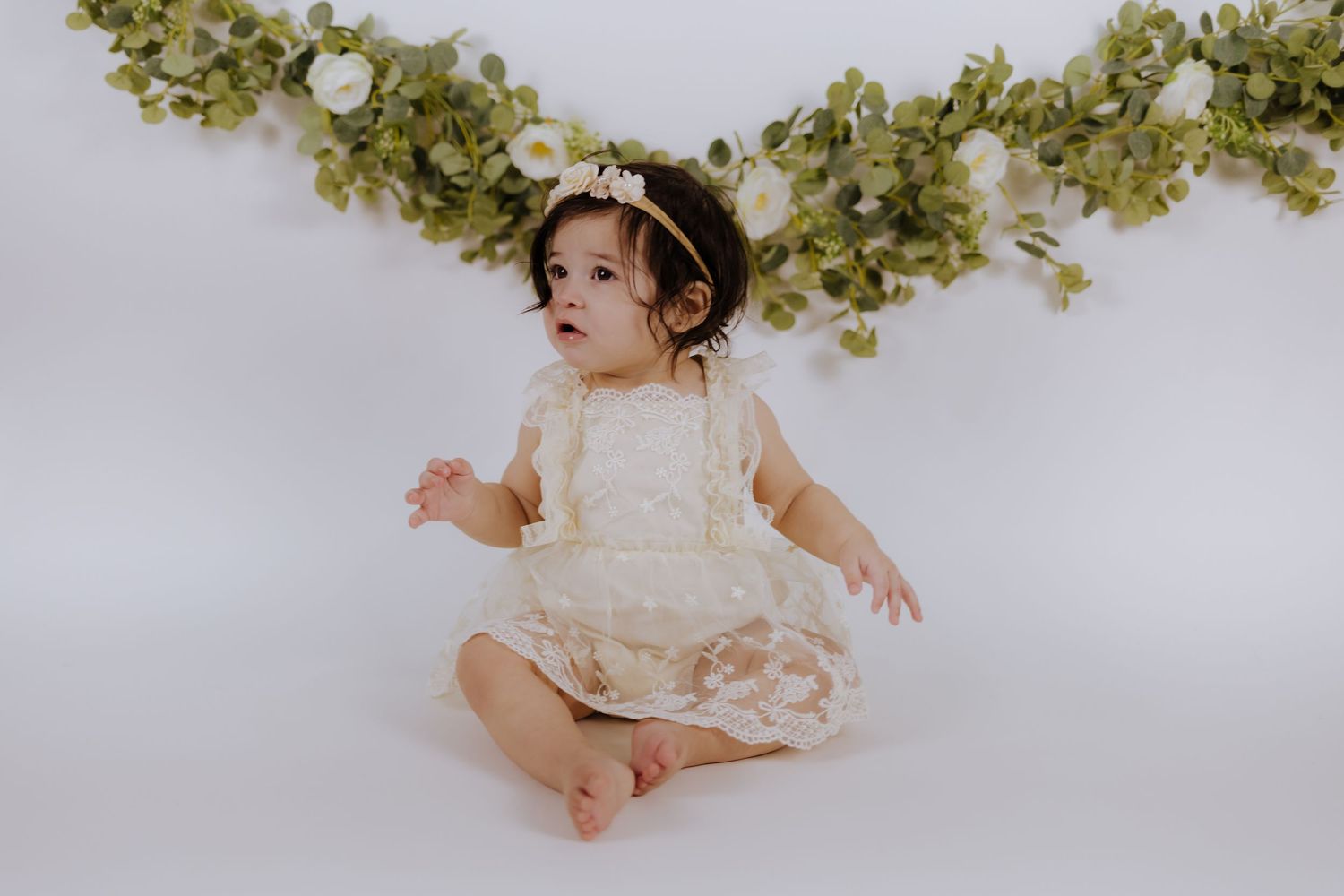 A baby girl in a cream lace romper and floral headband sits against a white backdrop with a green floral garland.