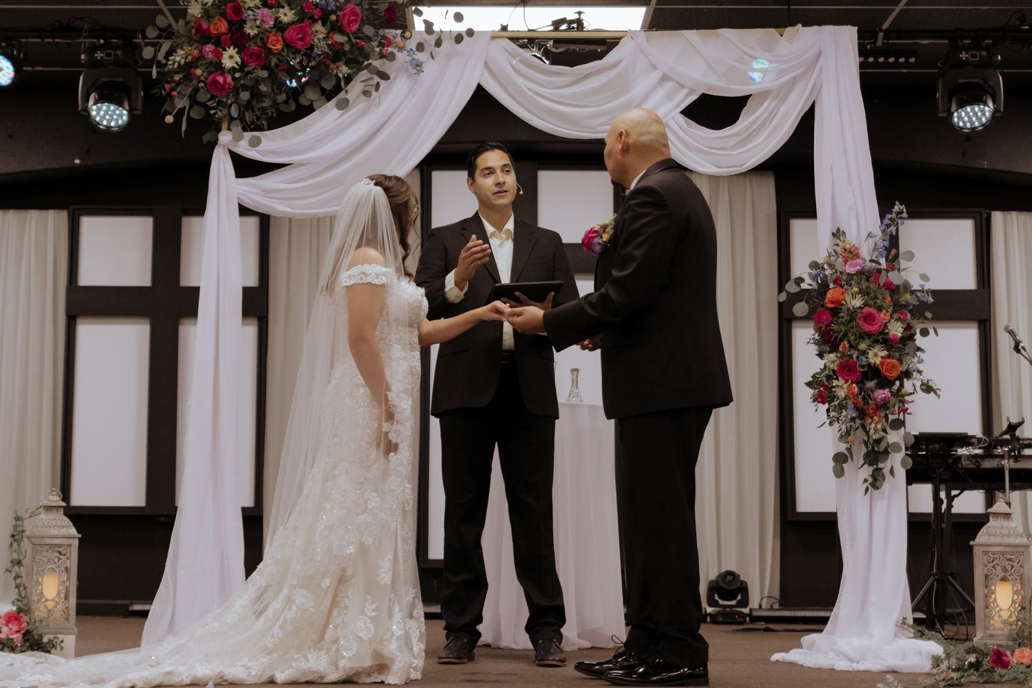 Couple exchanges vows at elegant indoor ceremony beneath white draped arch adorned with vibrant floral arrangements.